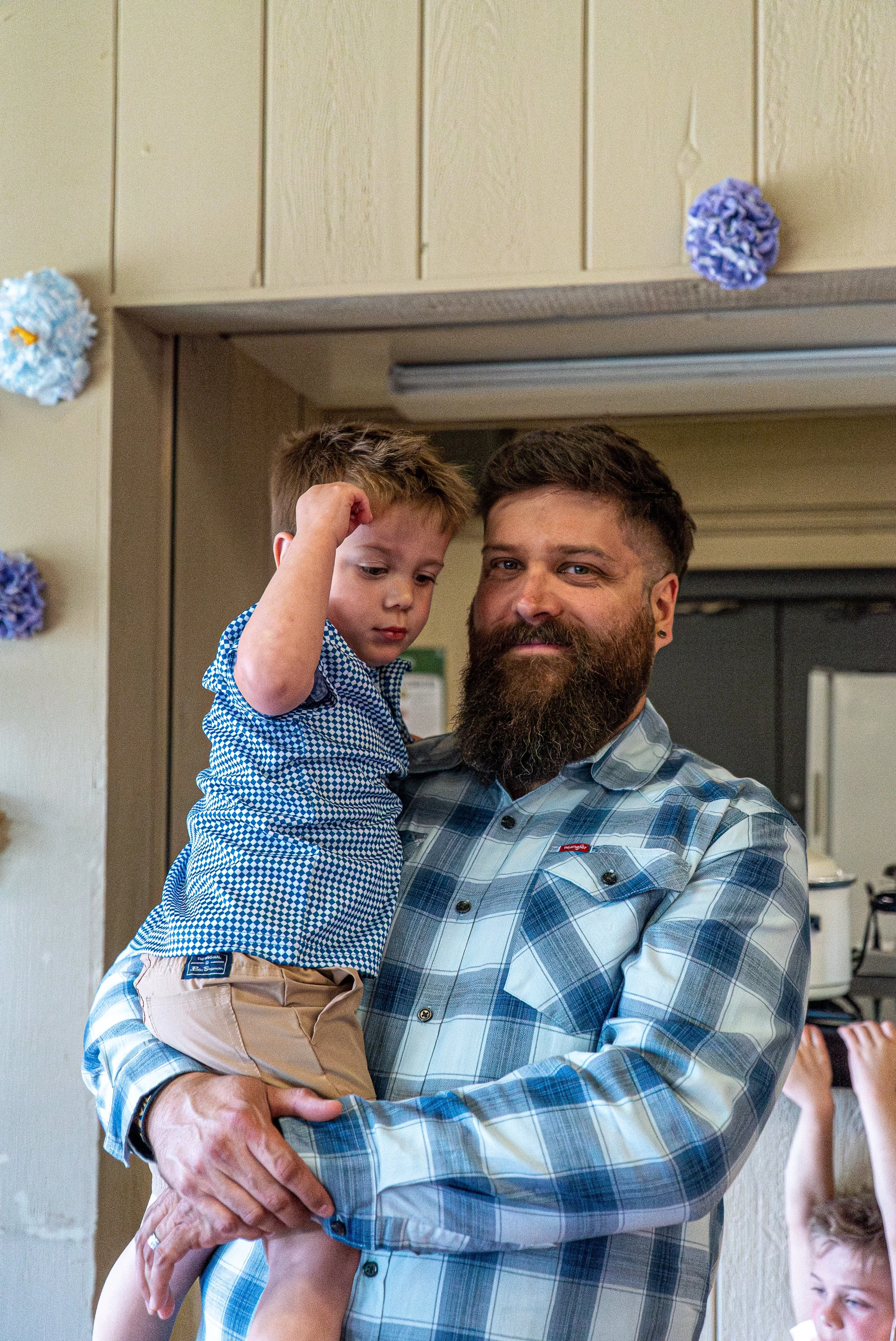 A bearded man with short dark hair wearing a blue plaid shirt holding a young boy with short blonde hair in his arms. The boy is wearing a blue patterned shirt and beige shorts, looking down with a serious expression. The background shows a beige wal