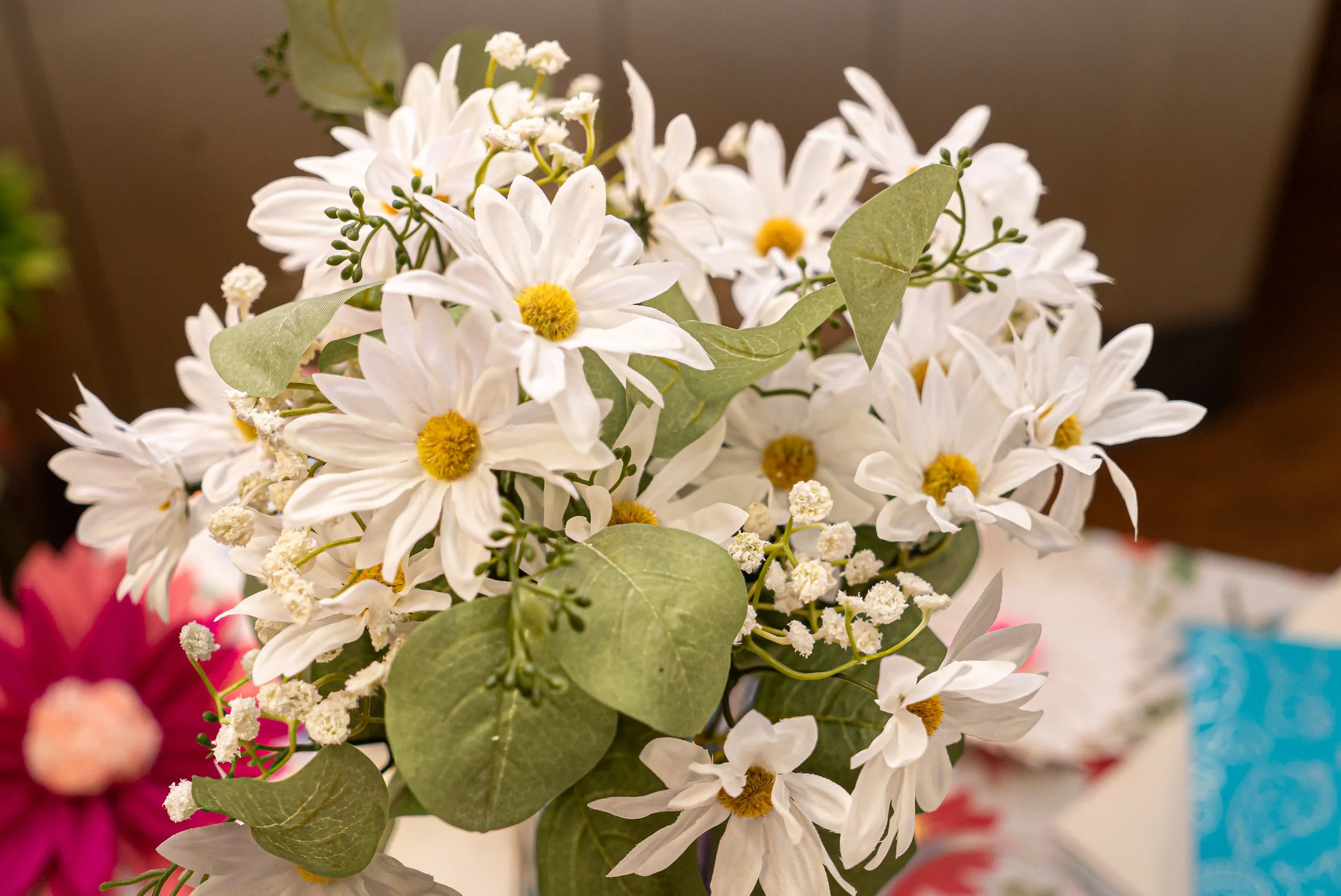 A bouquet of white daisies with yellow centers, green leaves, and small white filler flowers against a blurred background.