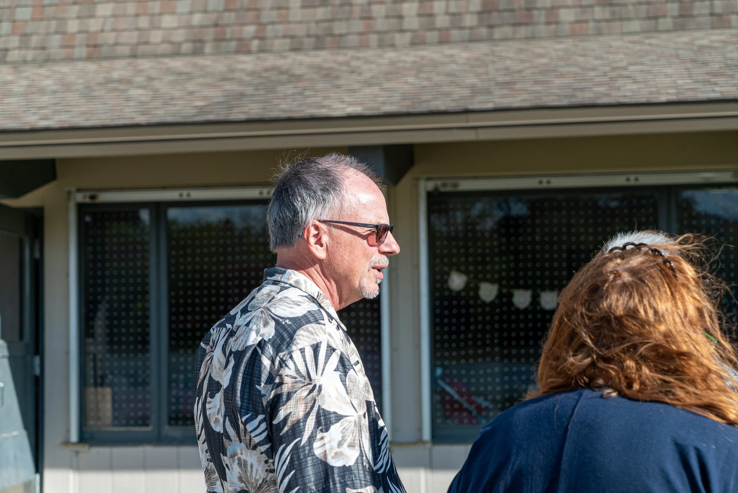 Side view of an elderly man wearing glasses and a patterned shirt, standing outdoors near a building with large window panes, with part of a woman with curly hair in the foreground.