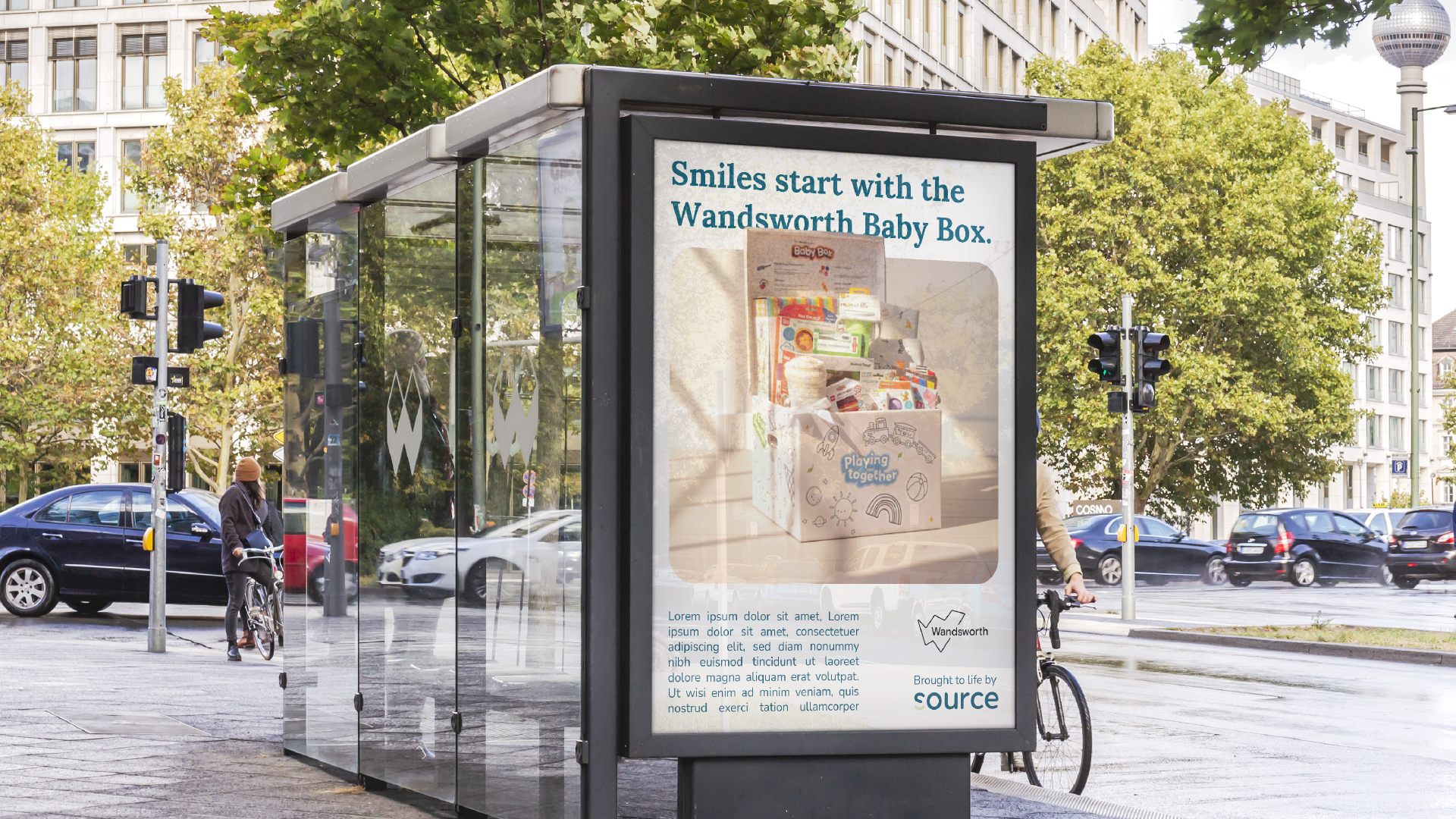 Bus stop shelter with an advertisement for Wandsworth Baby Box that says 'Smiles start with the Wandsworth Baby Box.' A bicycle is parked nearby, and a person walking a bike is visible in the background on a city street lined with trees and cars.