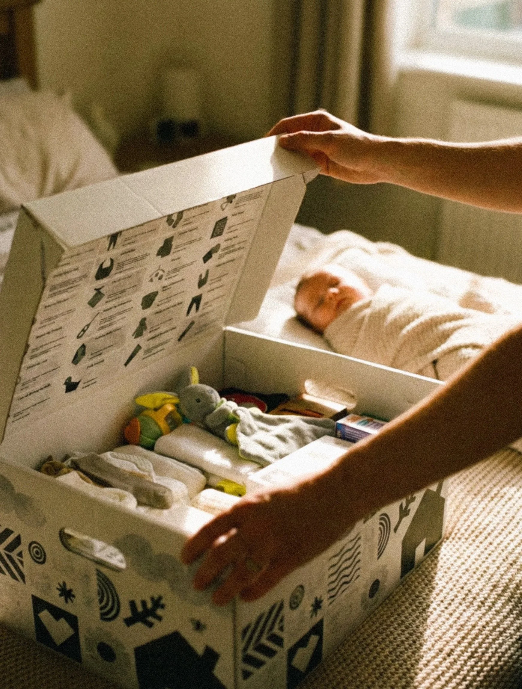 Person opening a box of baby essentials on a bed with a newborn baby wrapped in a blanket sleeping in the background.