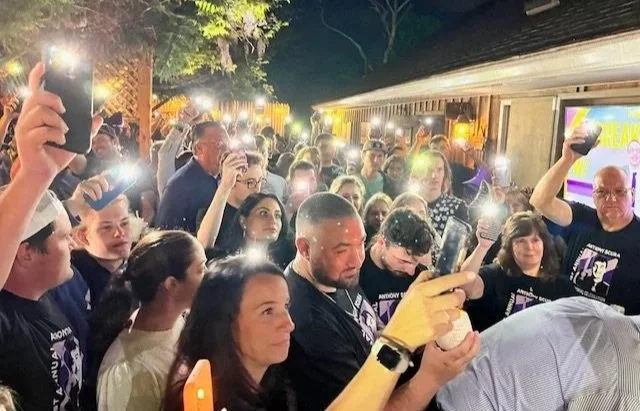 Crowd of people at an outdoor event taking photos with their phones at night, with some people wearing matching black t-shirts.
