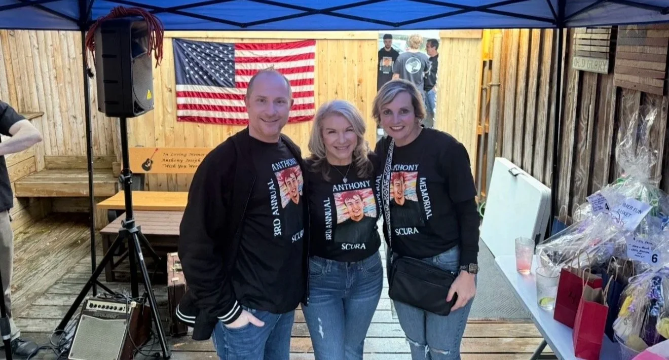 Three people wearing matching shirts with a caricature face design, standing in front of an American flag and a wooden fence, at an outdoor event with a blue canopy overhead.