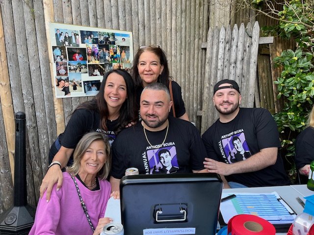 Five people gathered around a table outdoors, smiling for the camera, with a photo collage on a wooden fence in the background.