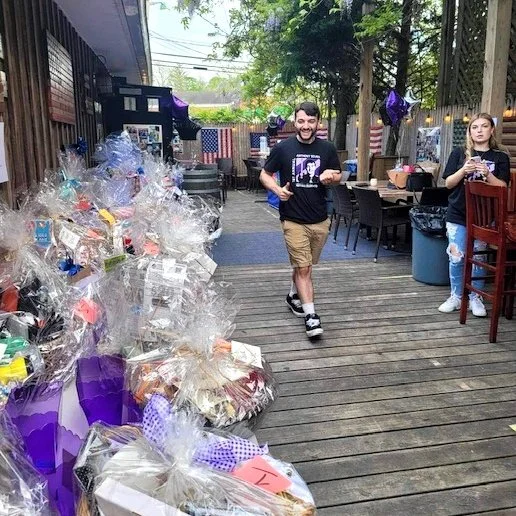 A man is walking on a wooden deck at an outdoor celebration, smiling and holding a cutout photo, with gift baskets wrapped in clear plastic along the side, and a woman in the background standing near tables decorated with balloons and American flags.