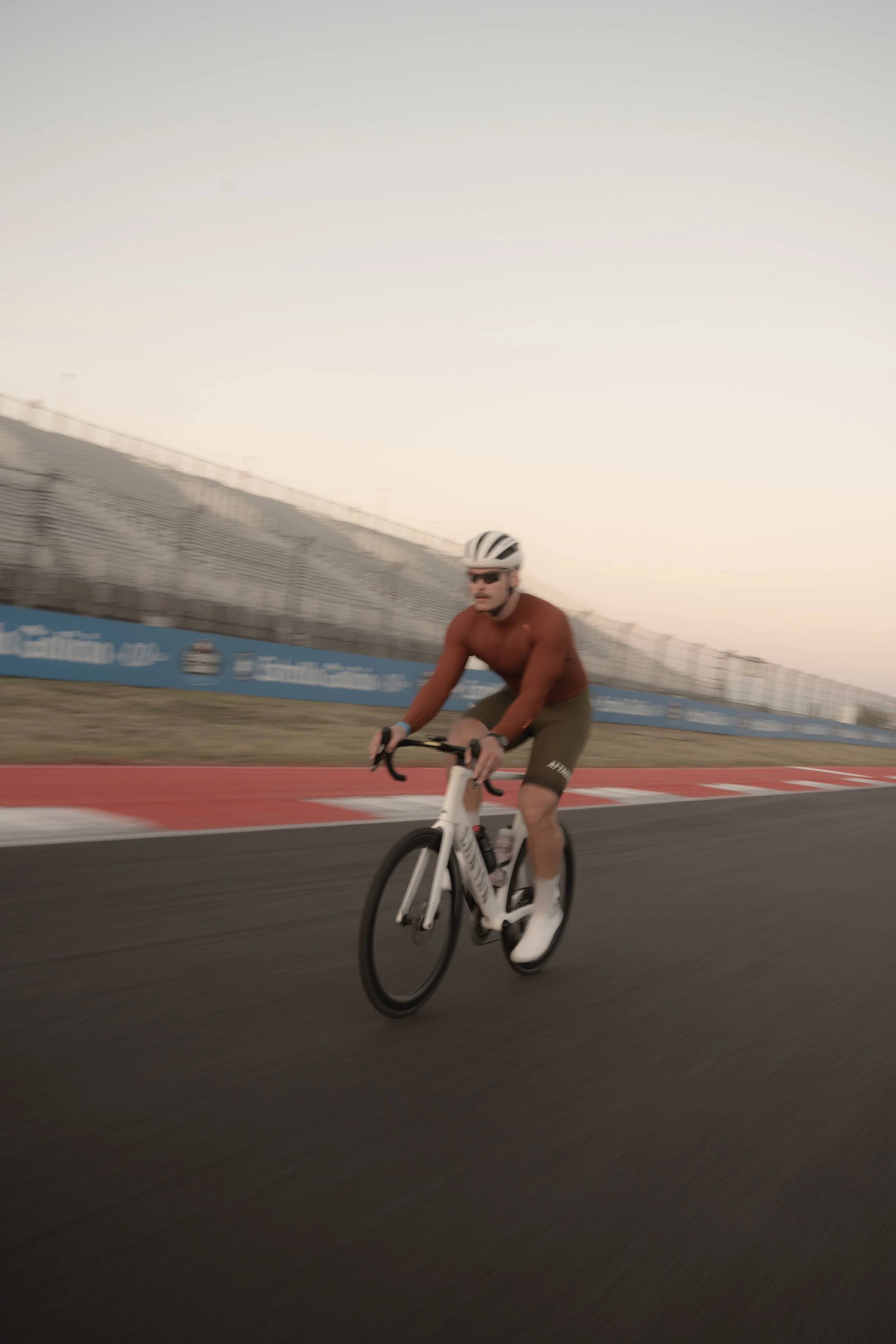 A man riding a white bicycle on a race track, wearing a helmet, sunglasses, a long-sleeve shirt, and shorts, with motion blur indicating high speed.