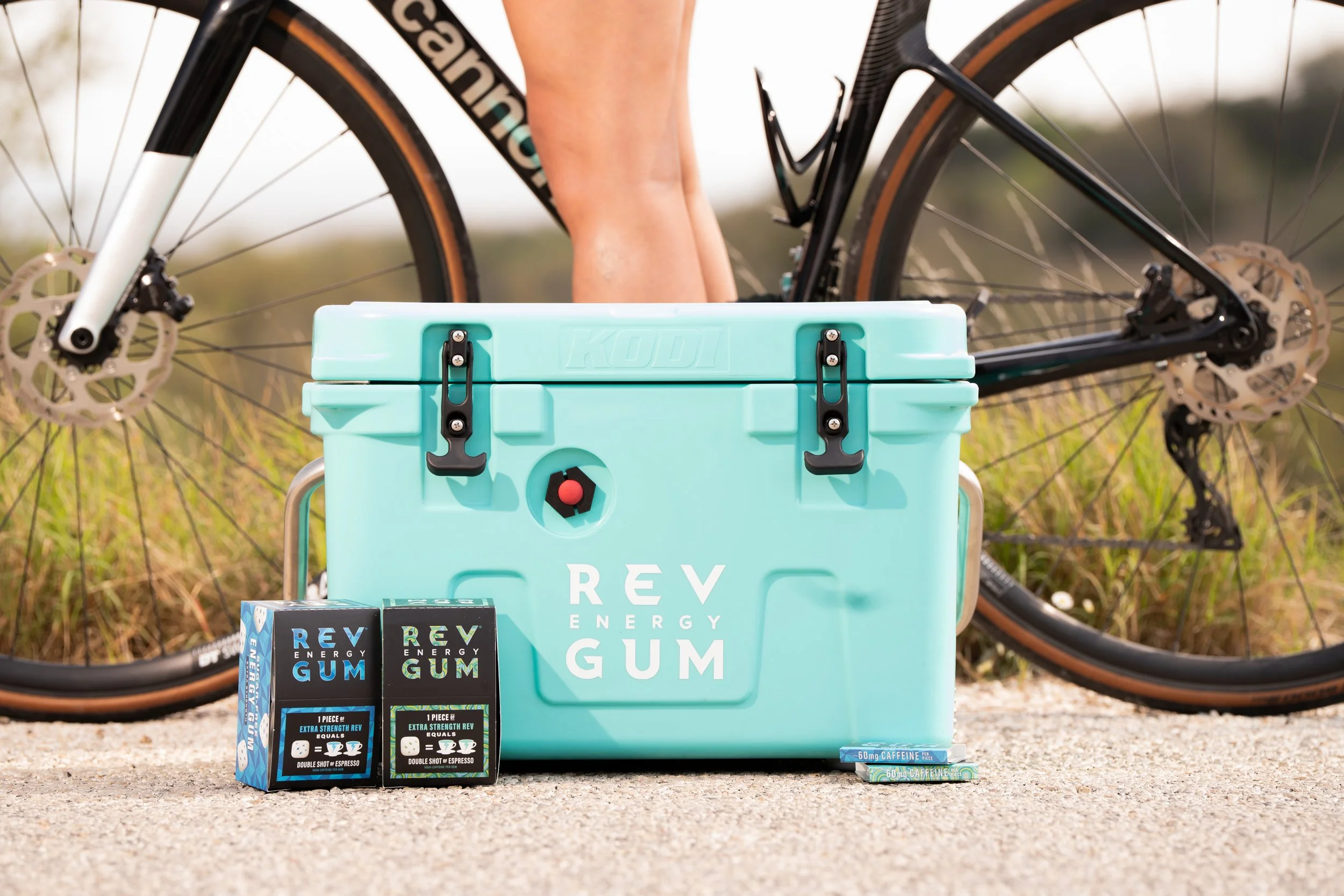 A turquoise cooler labeled 'REV ENERGY GUM' placed on the ground in front of a black mountain bike with disc brakes, with two packs of REV energy gum and a pack of caffeine gum beside it, on a concrete surface with grass and trees in the background.