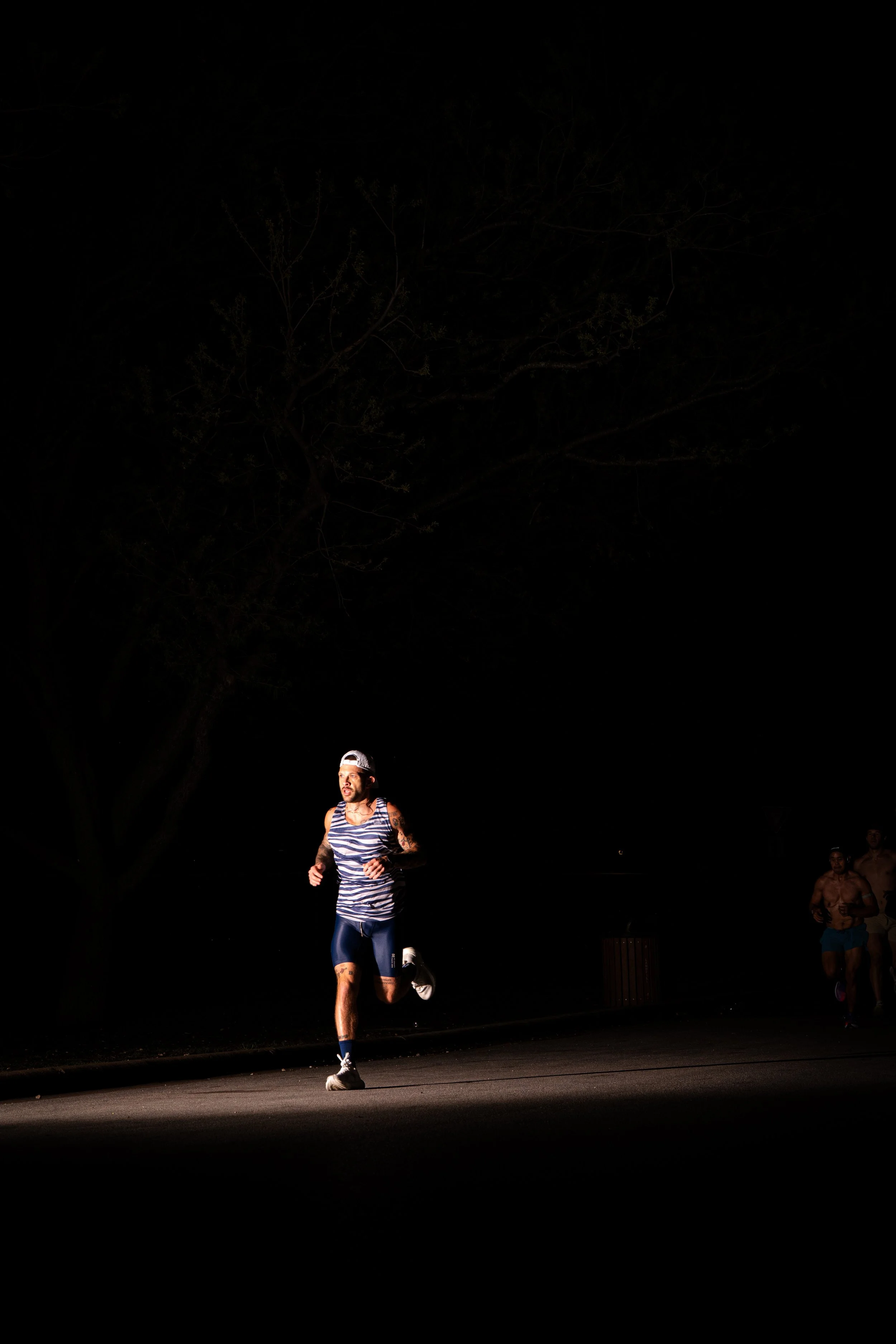 A man running at night on a dimly lit street, wearing a striped tank top, shorts, and a backwards cap, with other runners visible in the background.