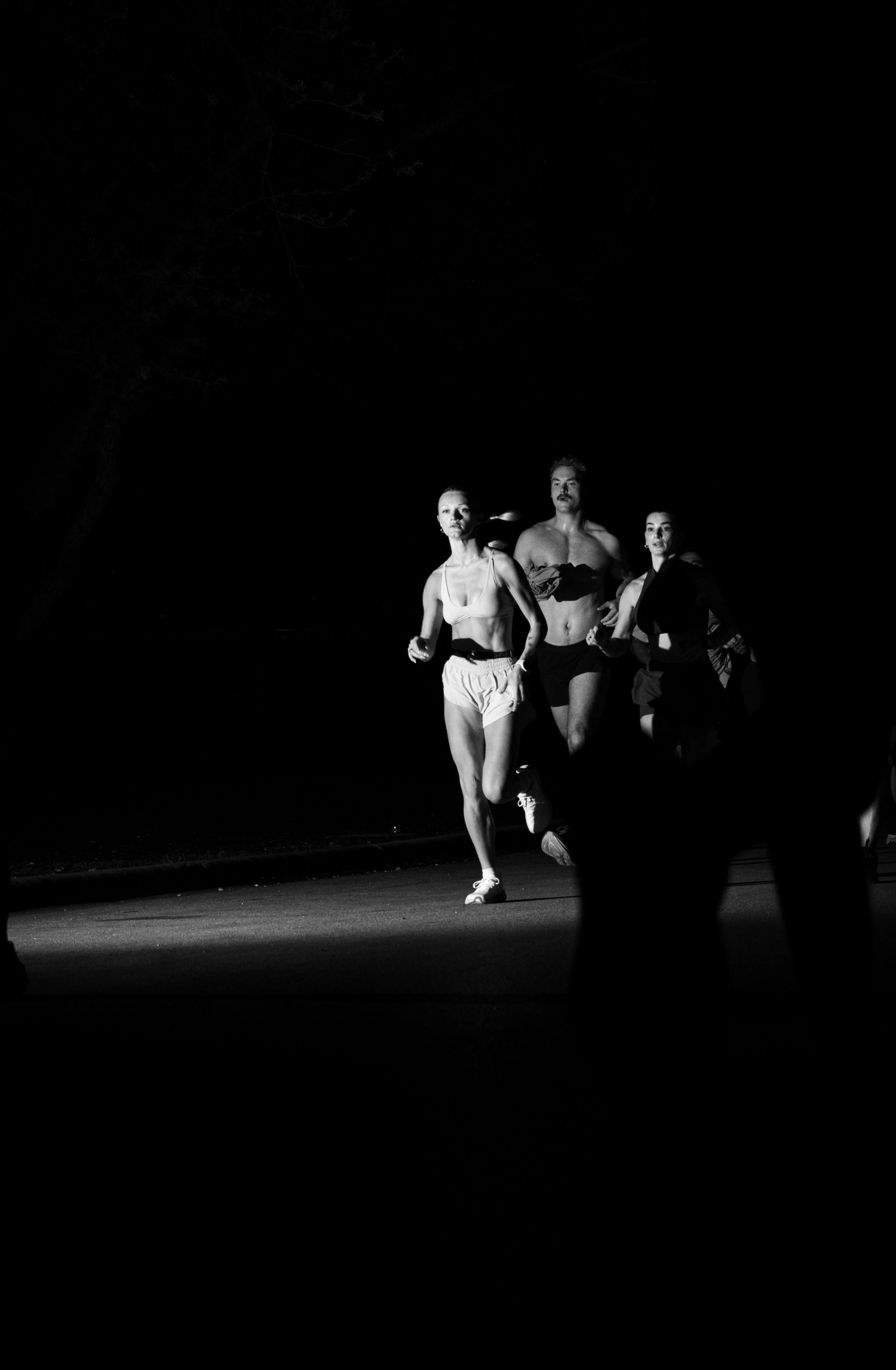Black and white photo of three runners, two women and one man, jogging outdoors at night, with trees in the background.