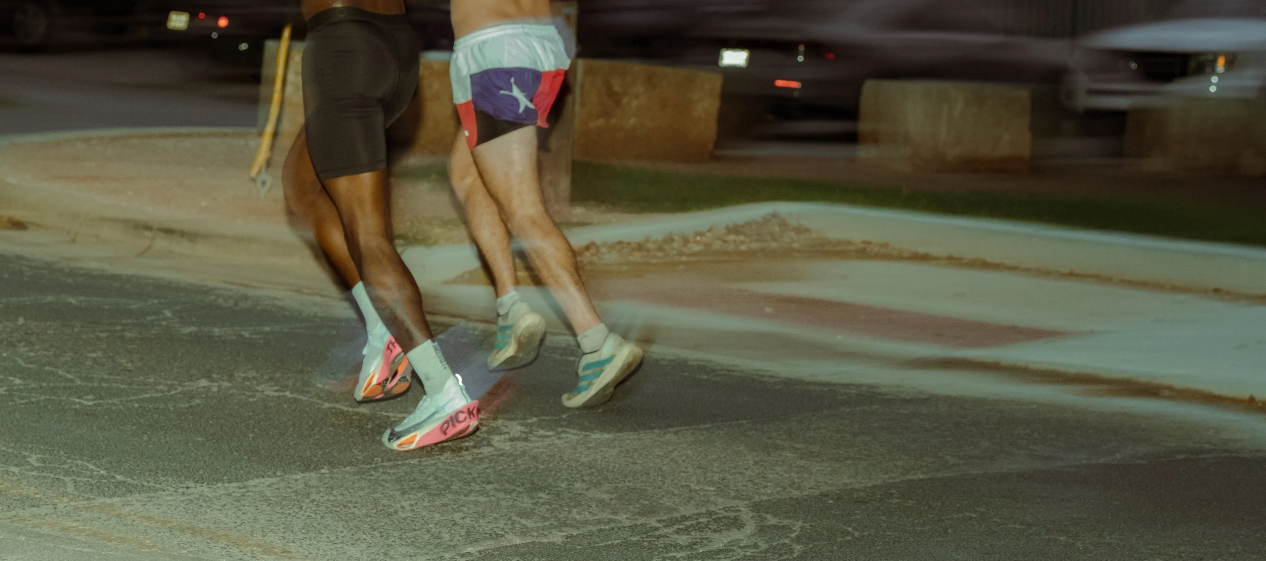 Two runners wearing shorts and running shoes running on a road at night with a blurry background.