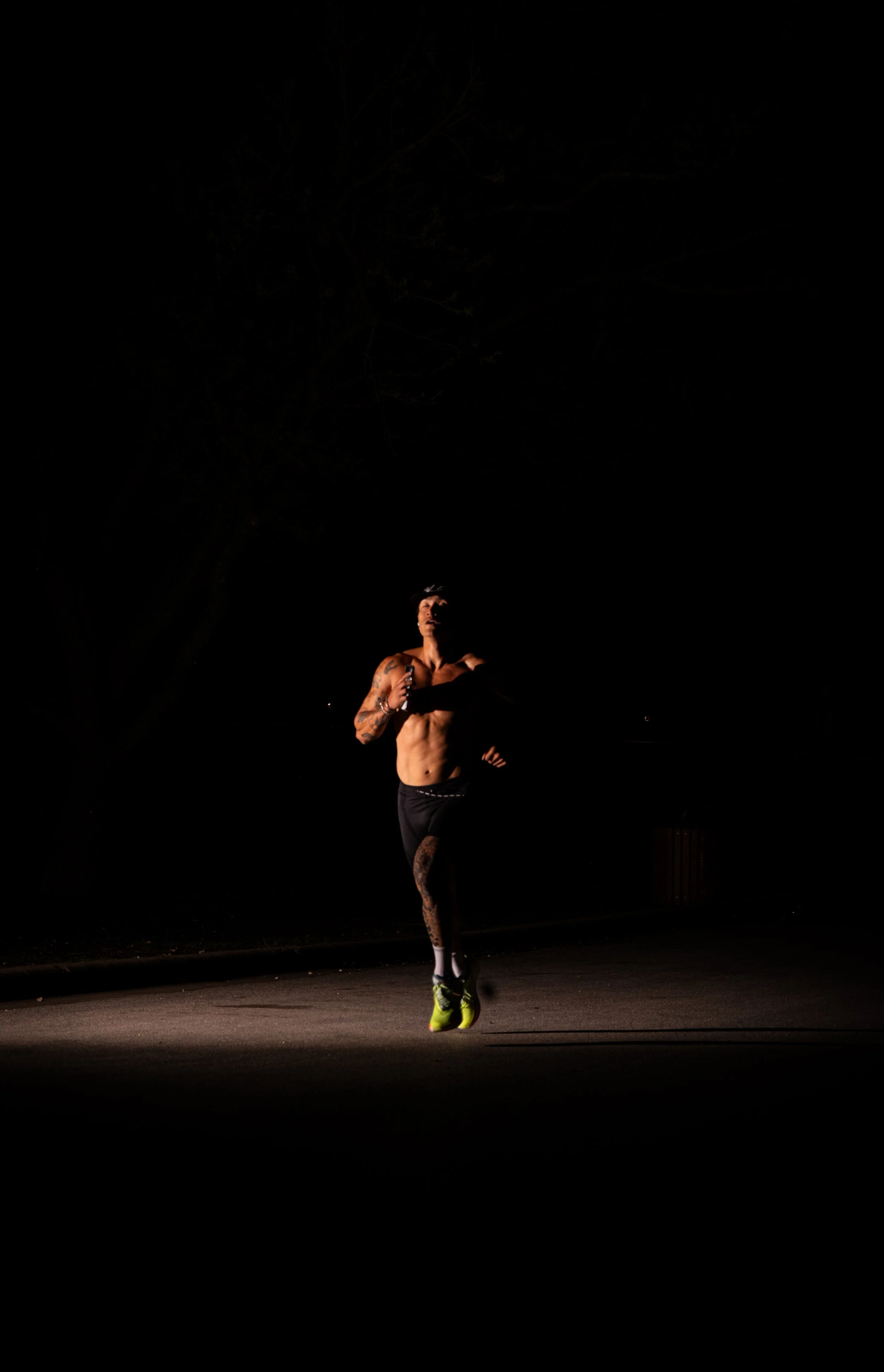 A male runner, shirtless and wearing black shorts, running at night under a dark sky with some trees in the background.