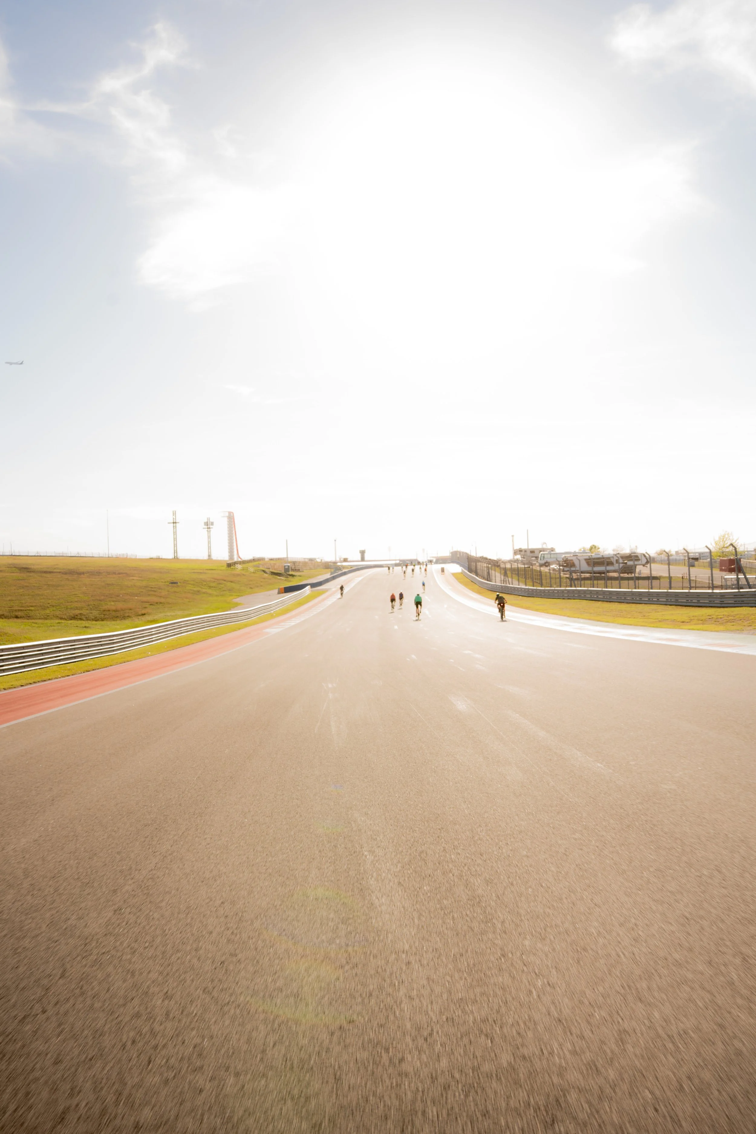 Race track with a few cyclists riding on it, fencing on the right side, and clear sky with clouds and bright sunlight.
