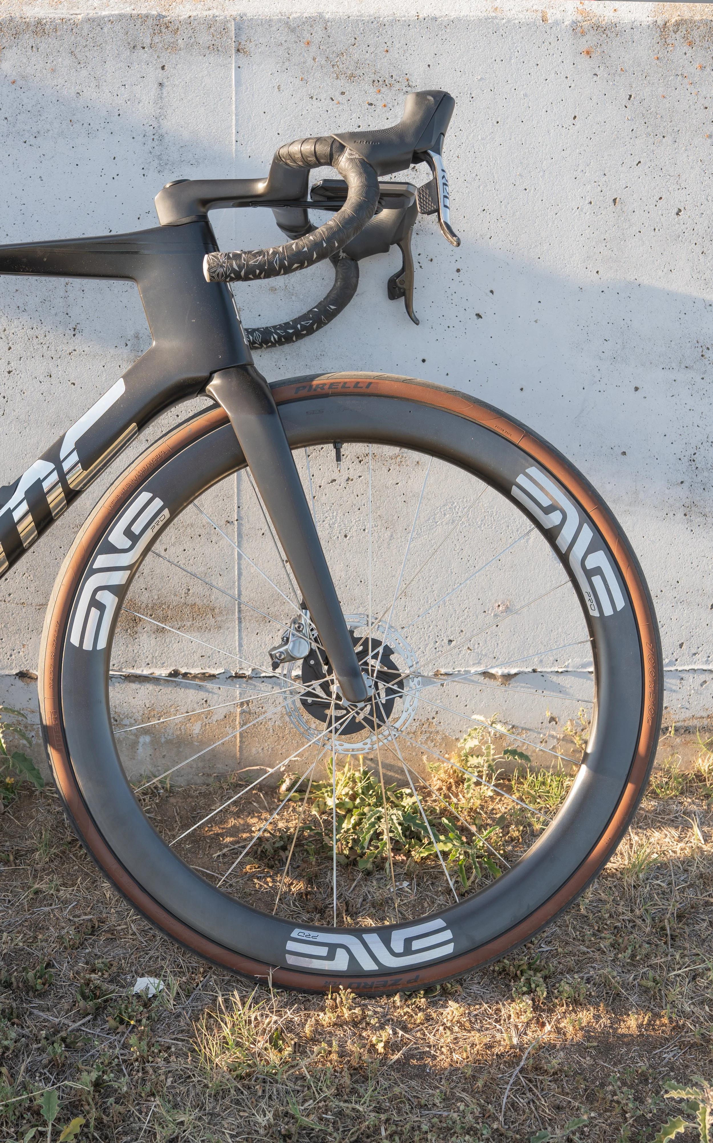 A black road bicycle with tan tires and aerodynamic design leaning against a white concrete wall, with some grass and weeds on the ground.