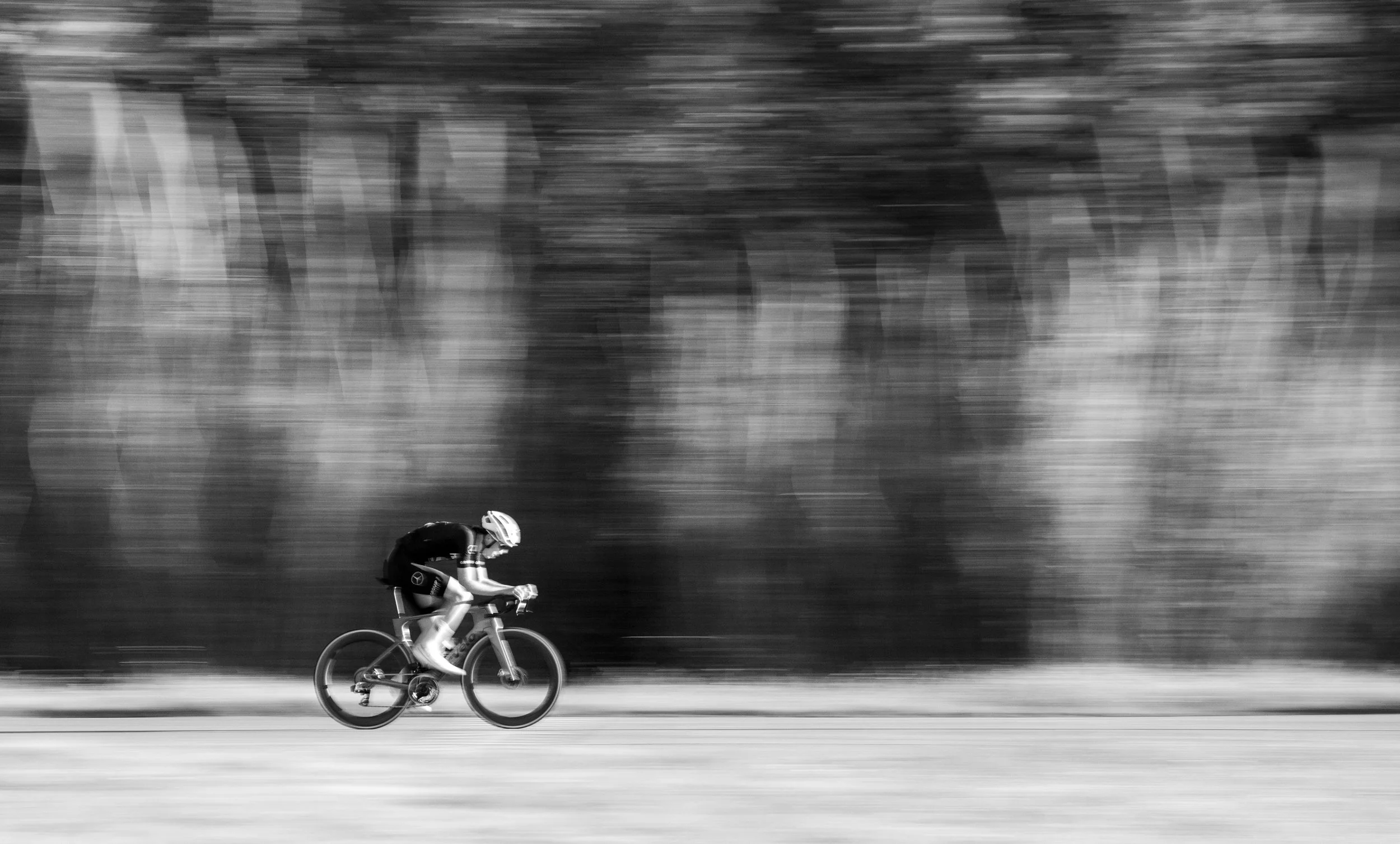 A person riding a bicycle on a road with a blurred natural background, in black and white.