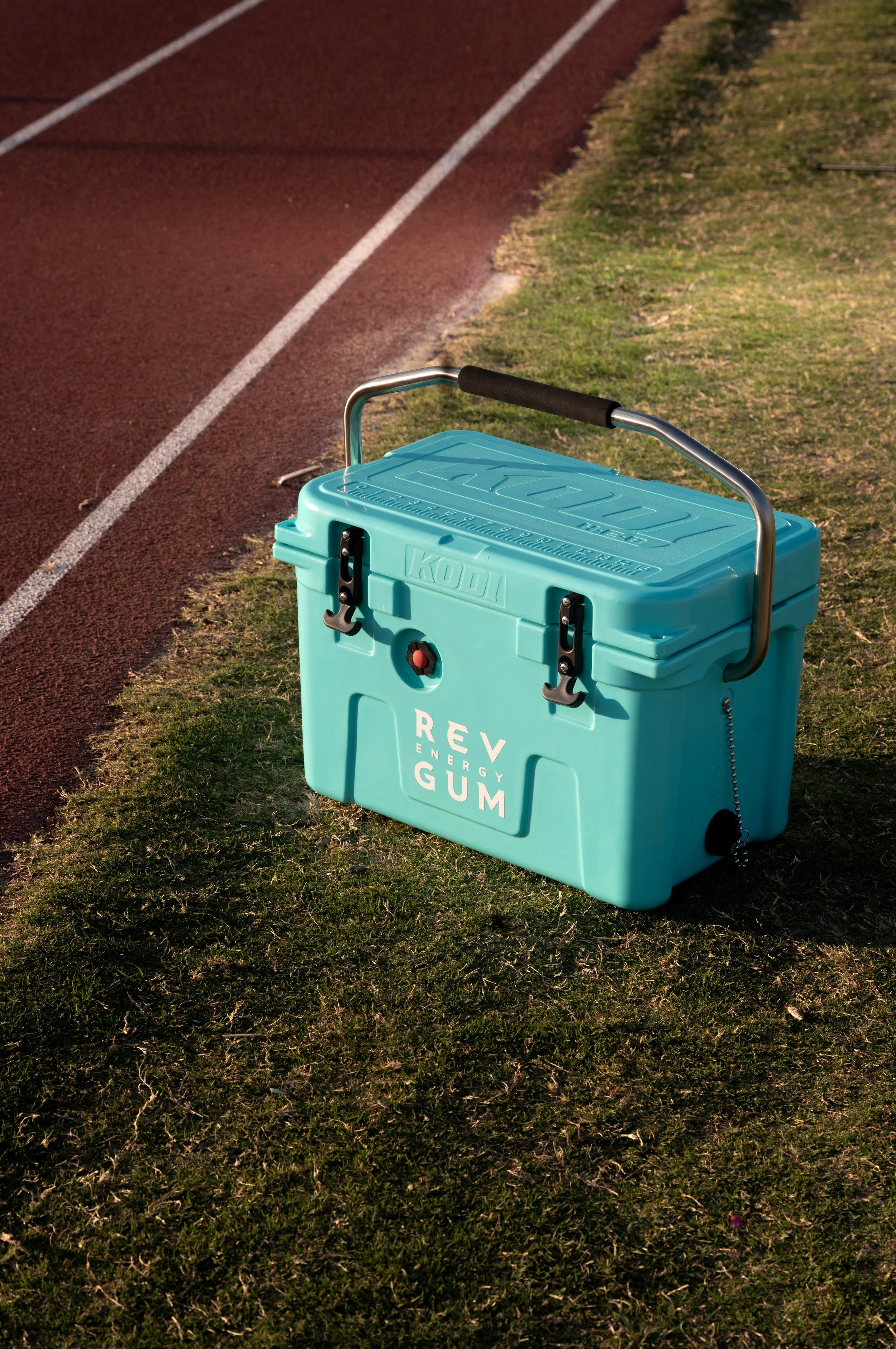 A teal cooler on a grassy area beside a running track, with the words 'REV ENERGY GUM' printed on it.