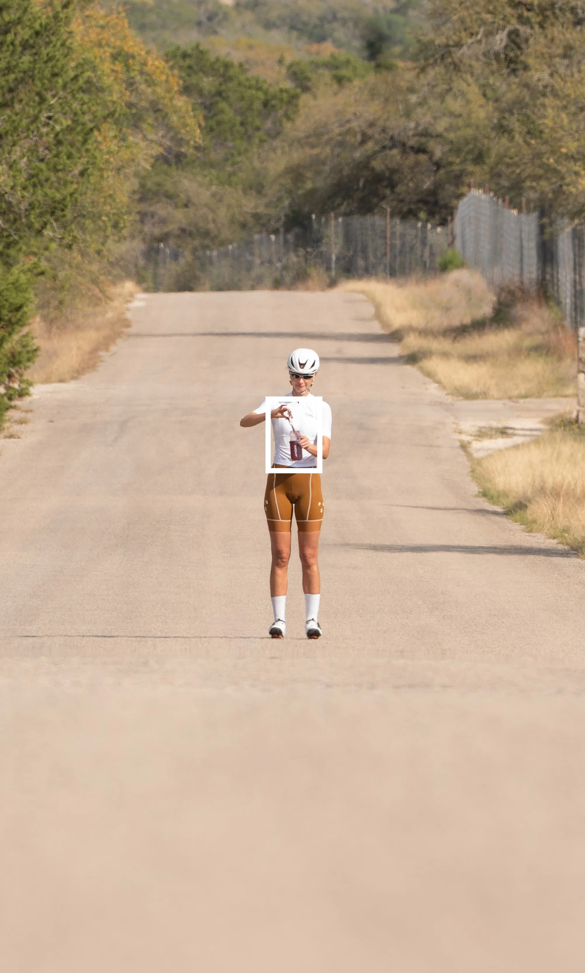 A cyclist standing on a deserted road adjusting their helmet and holding a water bottle, surrounded by trees and fencing.