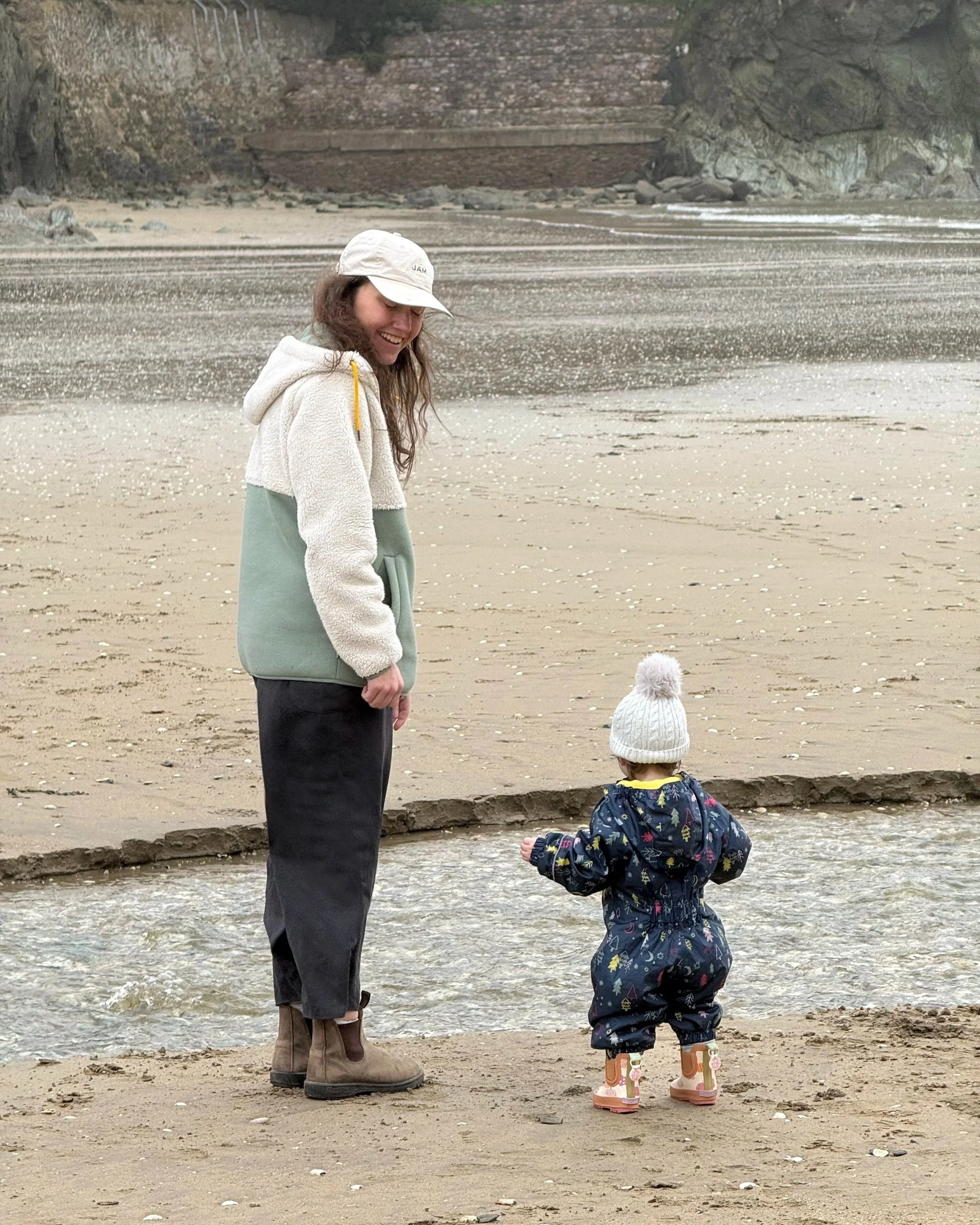 Alice standing on the beach in a fleece and baseball cap, next to her daughter in her waterproof onesie and wellie boots.