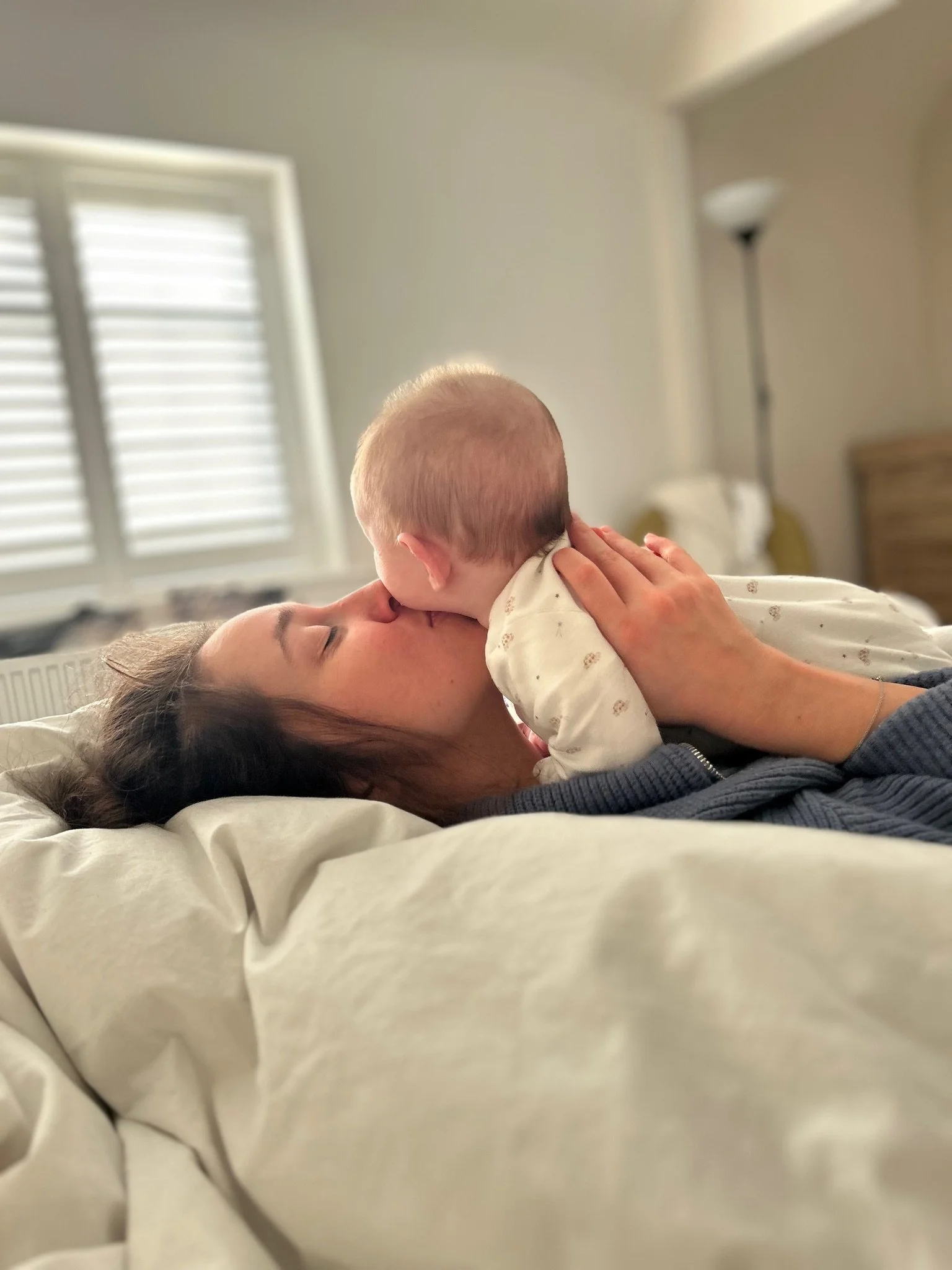 Alice lying on a bed holding her daughter close to her face, with sunlight coming through the shutters in the background.