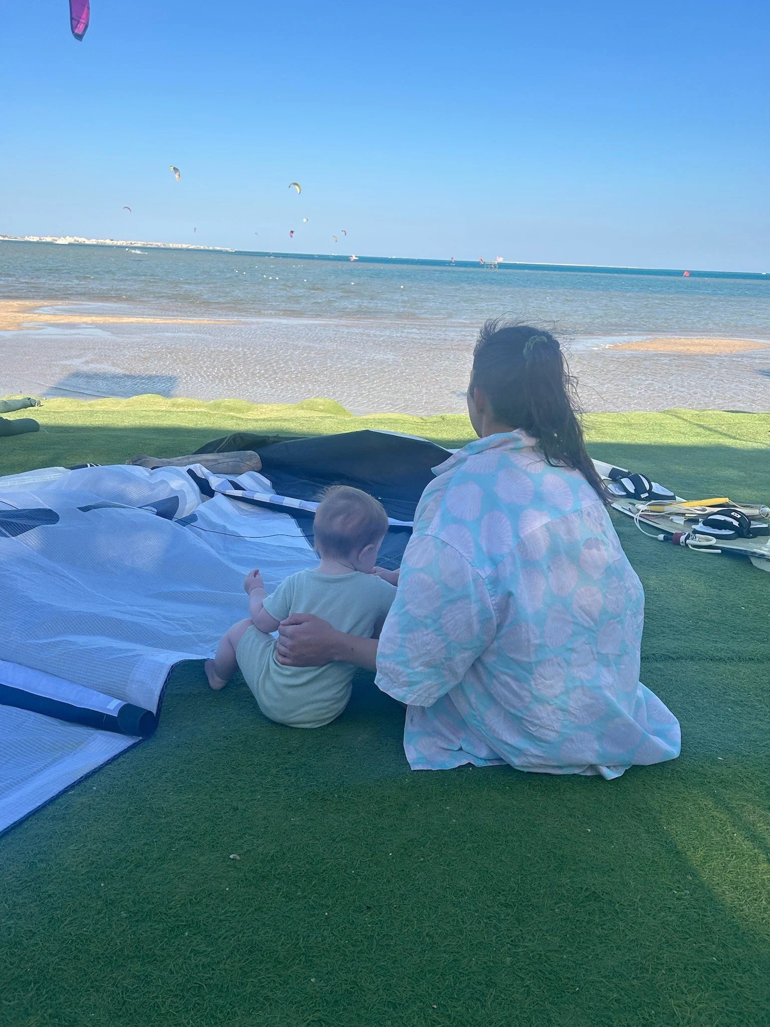 Alice sitting on the beach front next to a kite and her surfboard, with her daughter next to her.