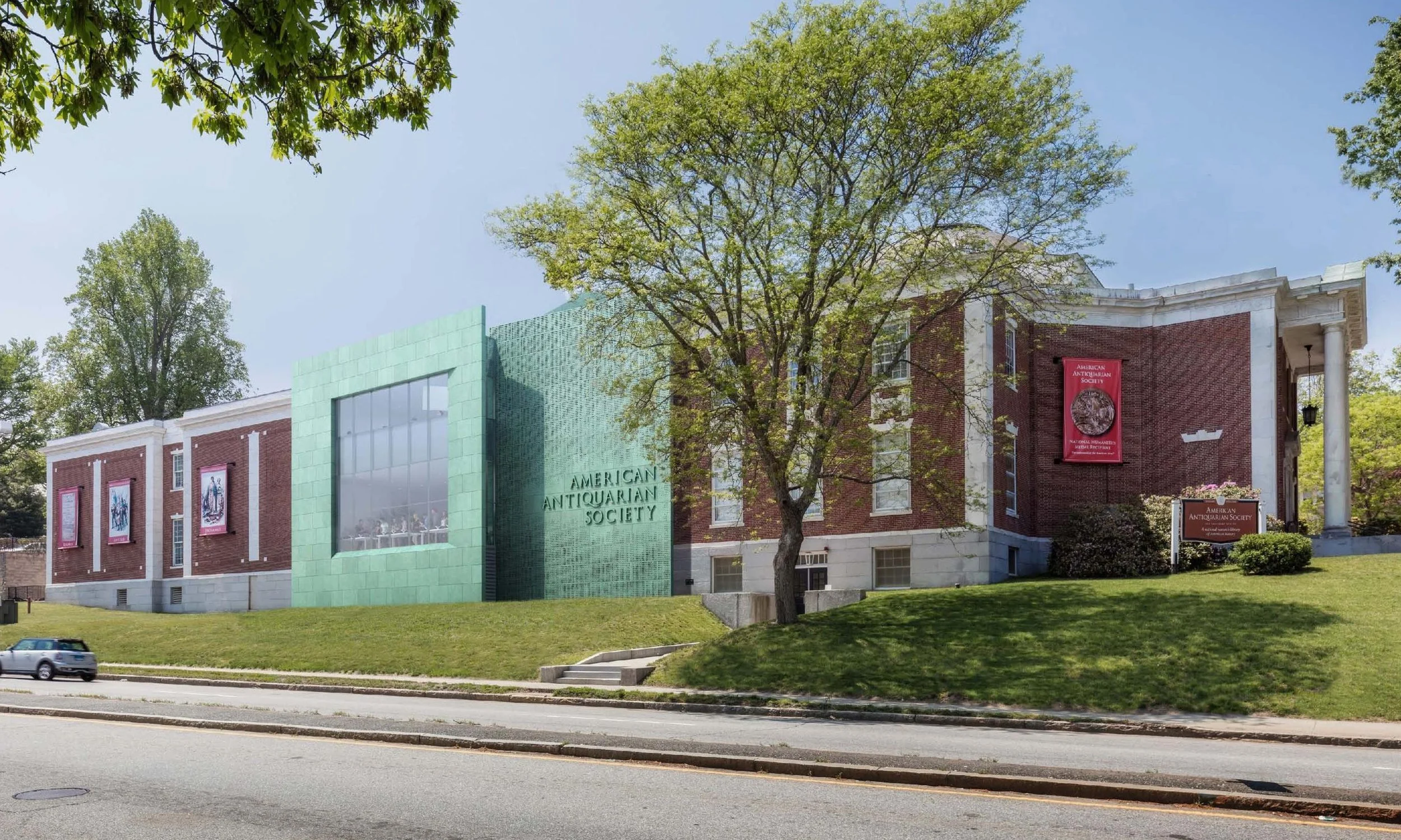 Building with red brick and white accents, displaying banners for the American Antiquarian Society, with a green modern extension on one side, surrounded by green trees and a grassy area, with cars on the street in front.