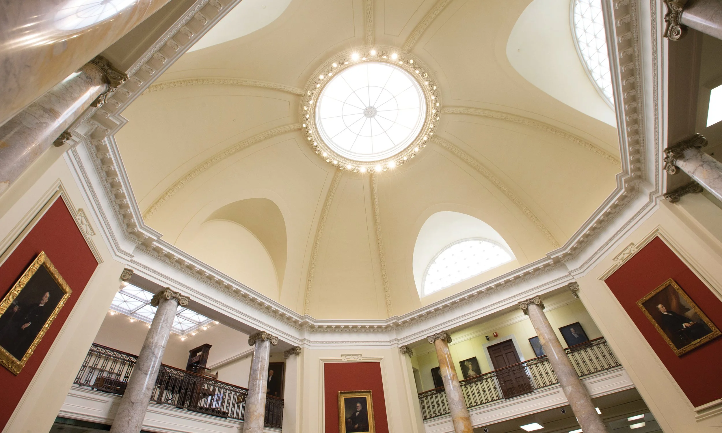 Interior view of a grand, historic building with a domed ceiling, large circular skylight, marble columns, and portraits on the walls. American Antiquarian Society, Worcester, Massachusetts