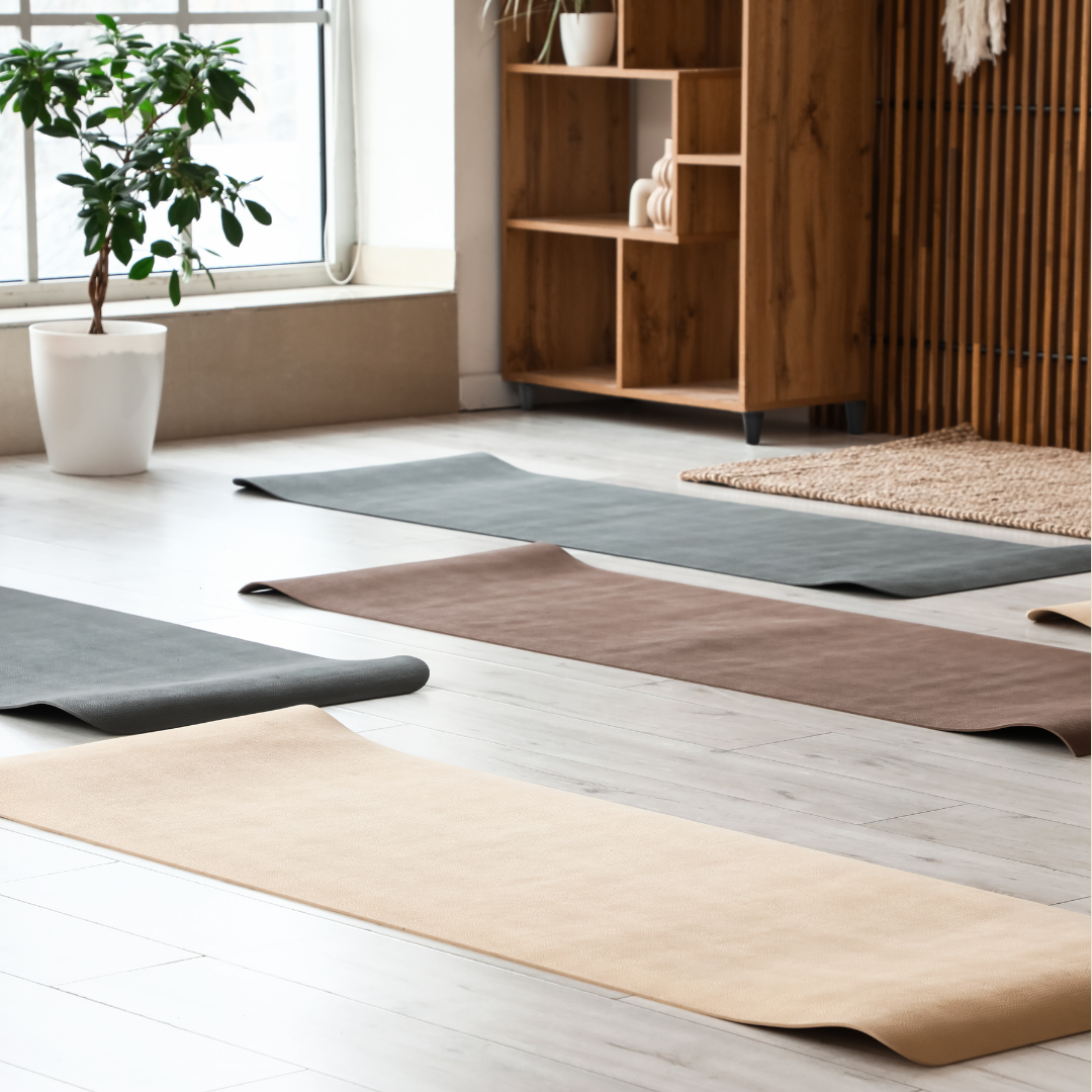 Indoor space with four yoga mats in different neutral colors laid out on light wood flooring, near a large window, a potted plant, and a wooden shelf with decorative items.