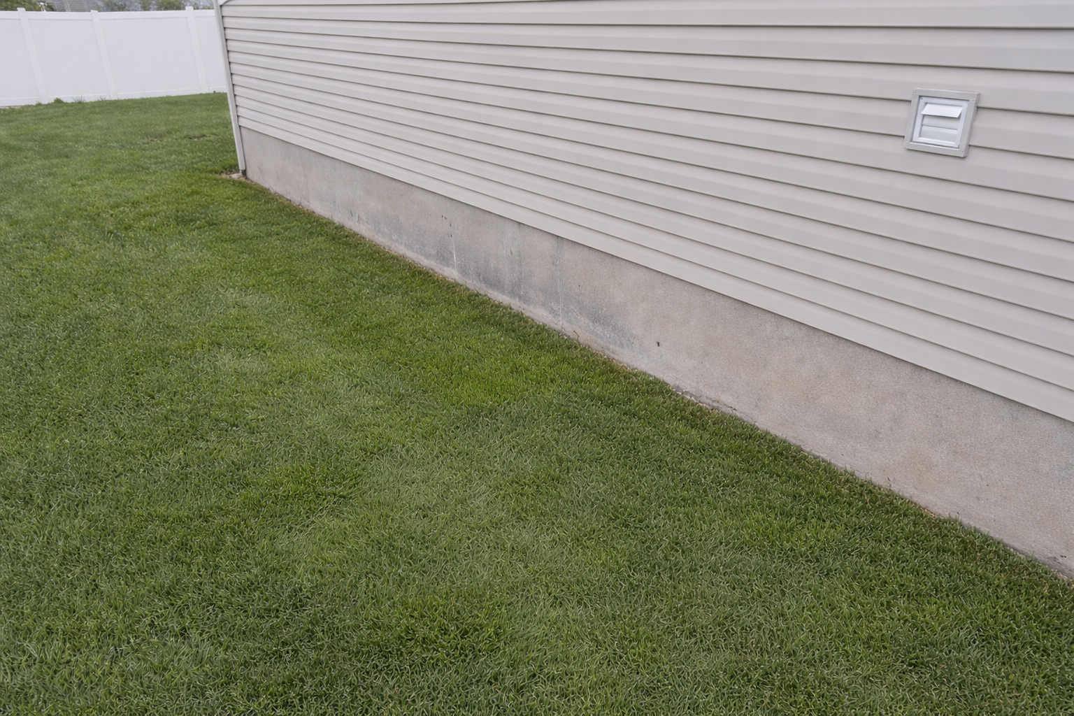 Close-up view of the beige siding and concrete foundation of a house, with a small vent on the siding, and green grass in the foreground.