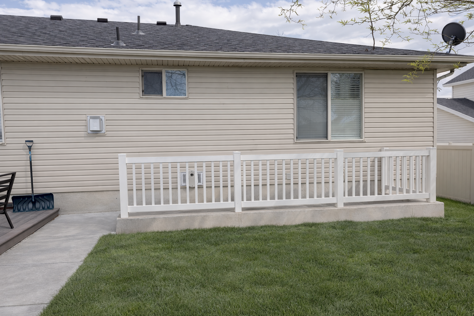 Backyard of a house with a small wooden deck, a white fence, and a grassy lawn. The house has beige siding, two windows, and a satellite dish on the roof.