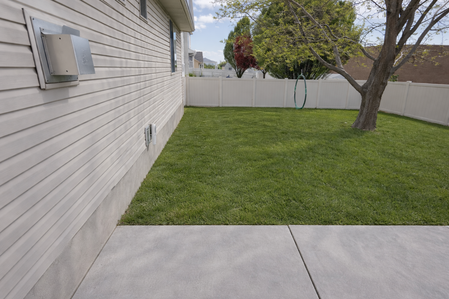 A backyard with a green lawn, a large tree, a white fence, a hanging hoop, and a side of a house with a beige vinyl siding wall, electrical outlets, and an electrical panel.