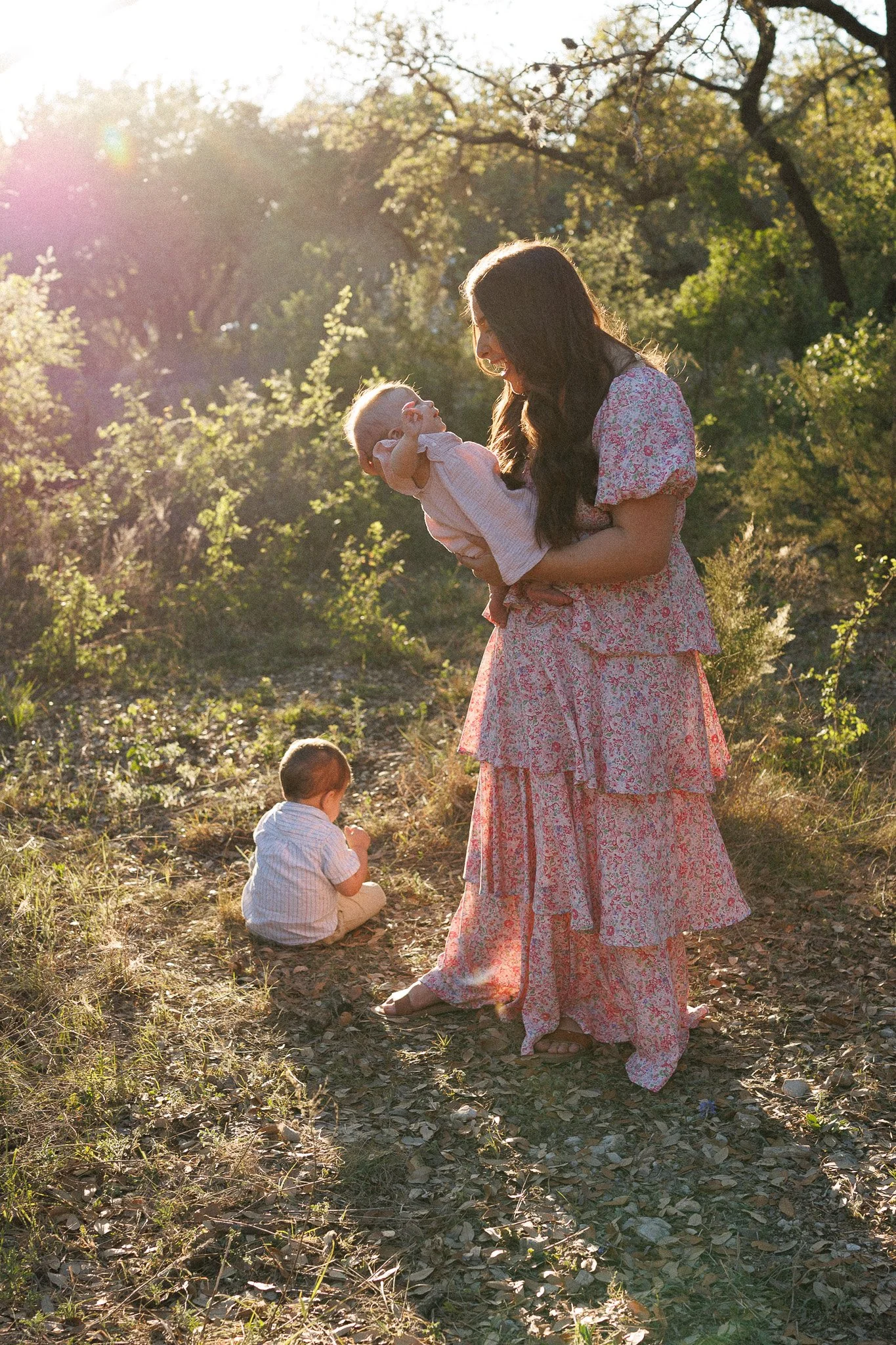 Woman in pink floral dress holding a smiling baby girl up in a sunny outdoor wooded area, with another young child sitting on the ground nearby.