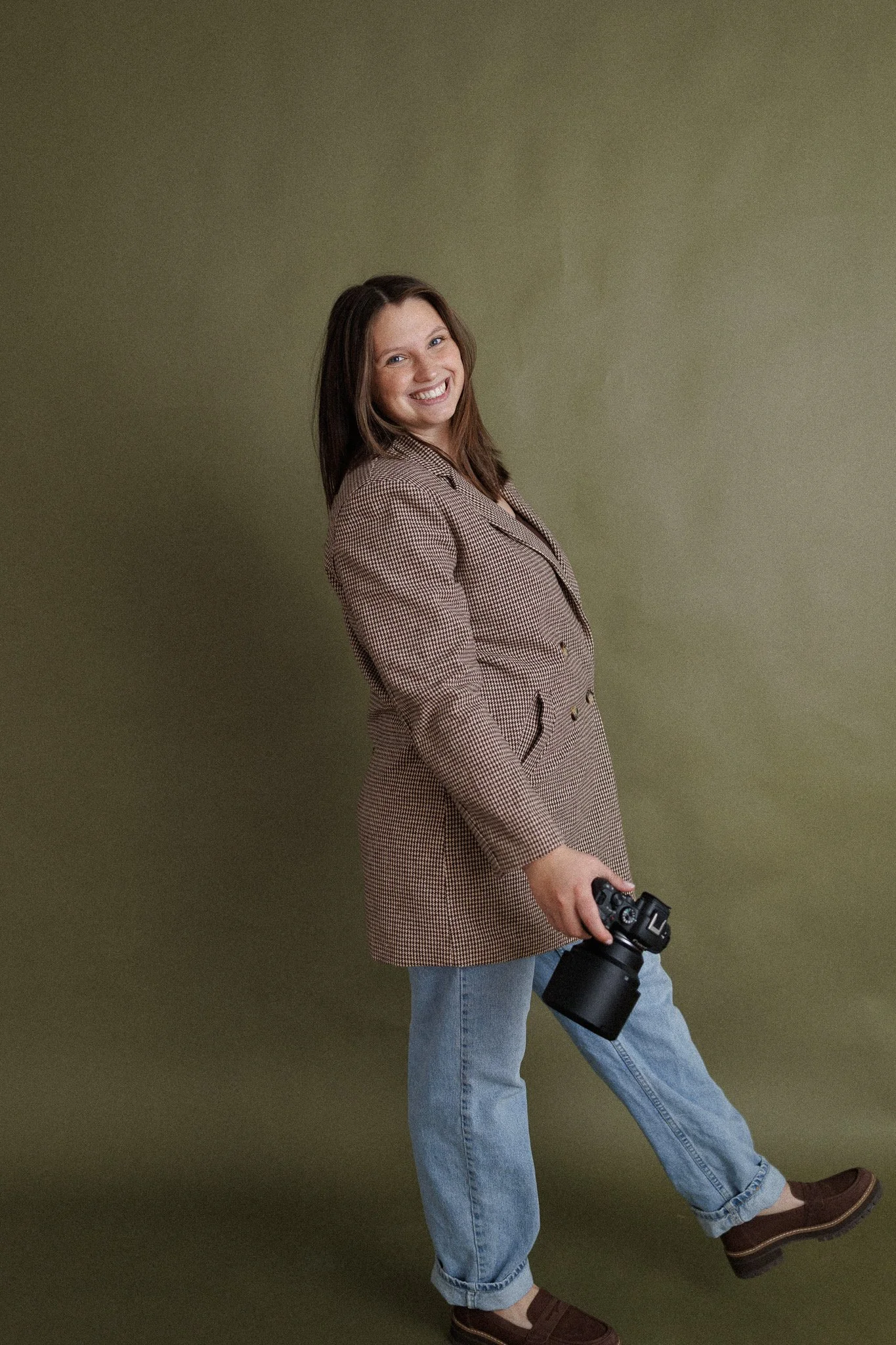 A woman with long dark hair, wearing a brown checked blazer and blue jeans, standing on a green background, smiling, holding a camera in her right hand.
