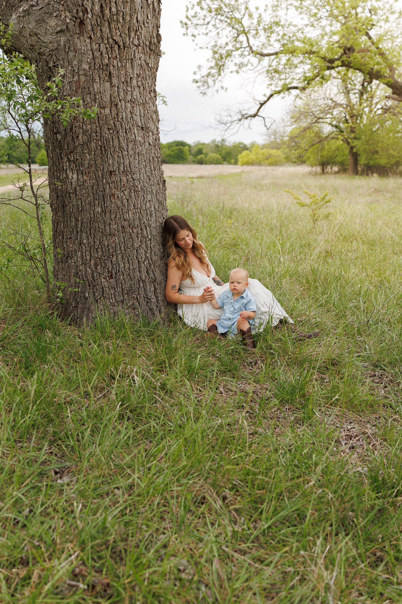 A woman sitting under a large tree holding hands with a young boy in a grassy field.