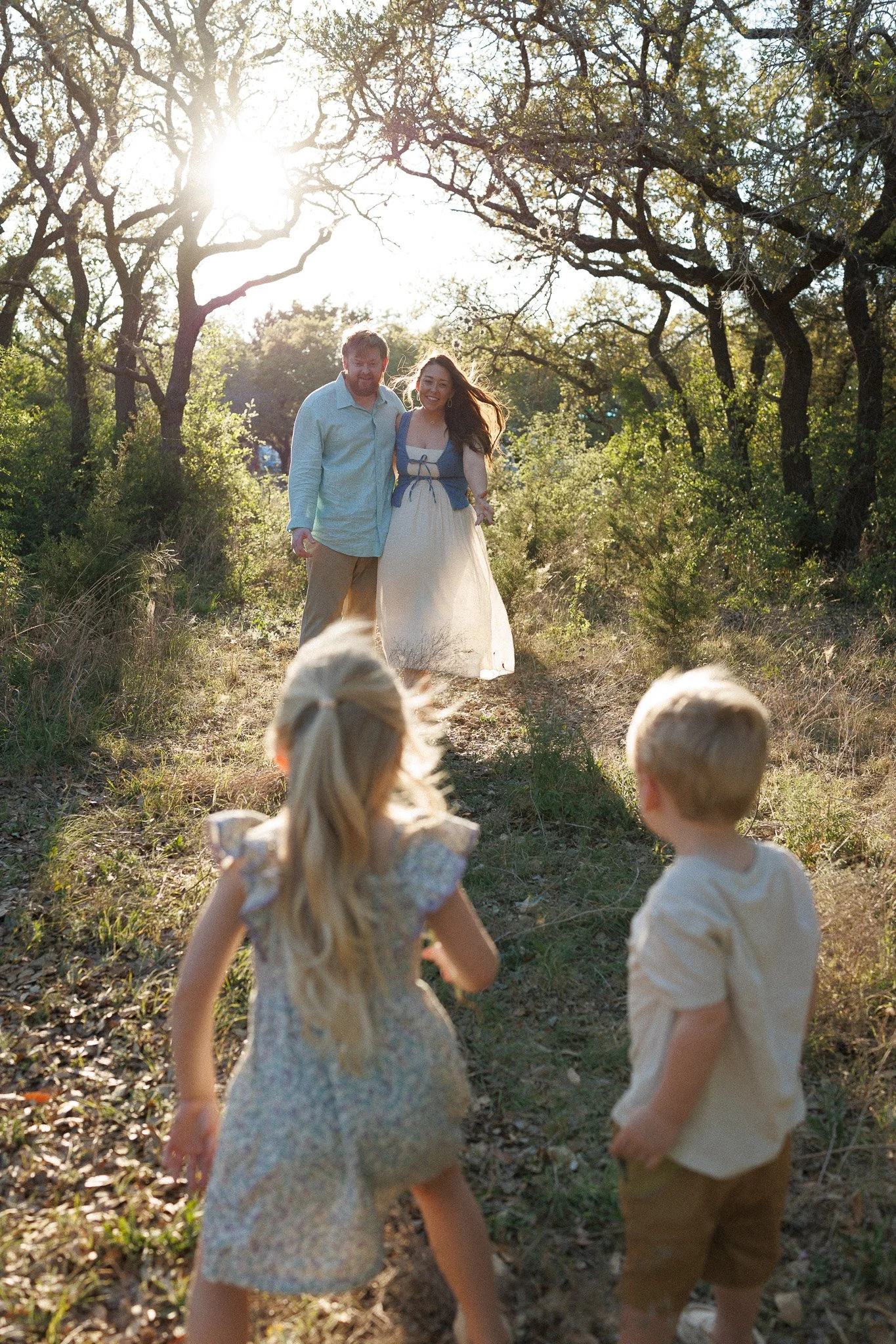 A family walking on a dirt trail in a wooded area during daytime. The sun is shining through the trees. In the background, a couple is walking towards the camera, smiling, while two young children are in the foreground, facing the couple.