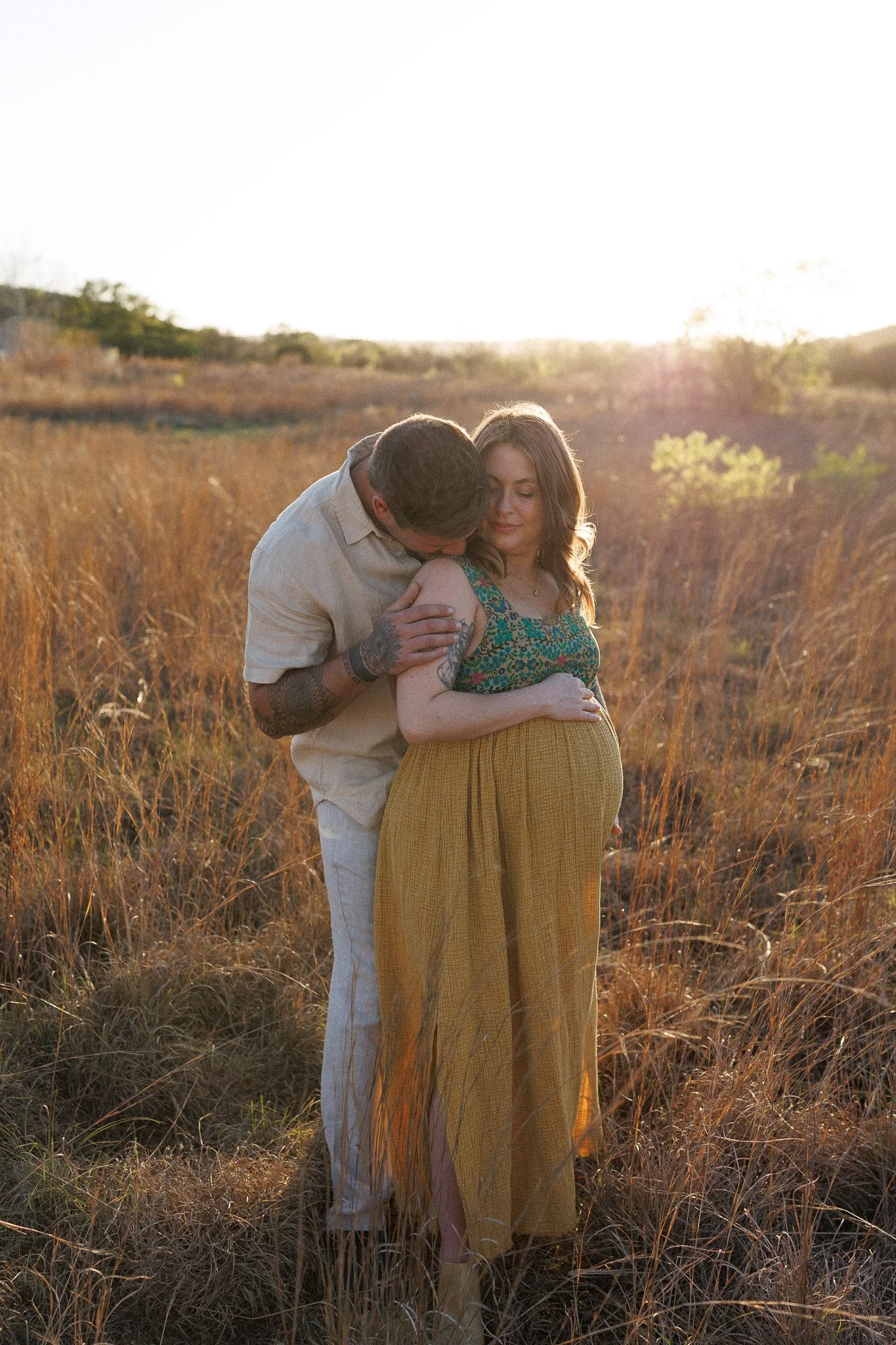 A pregnant woman in a floral top and yellow skirt standing in a grassy field with a man behind her, embracing her and kissing her shoulder during sunset.
