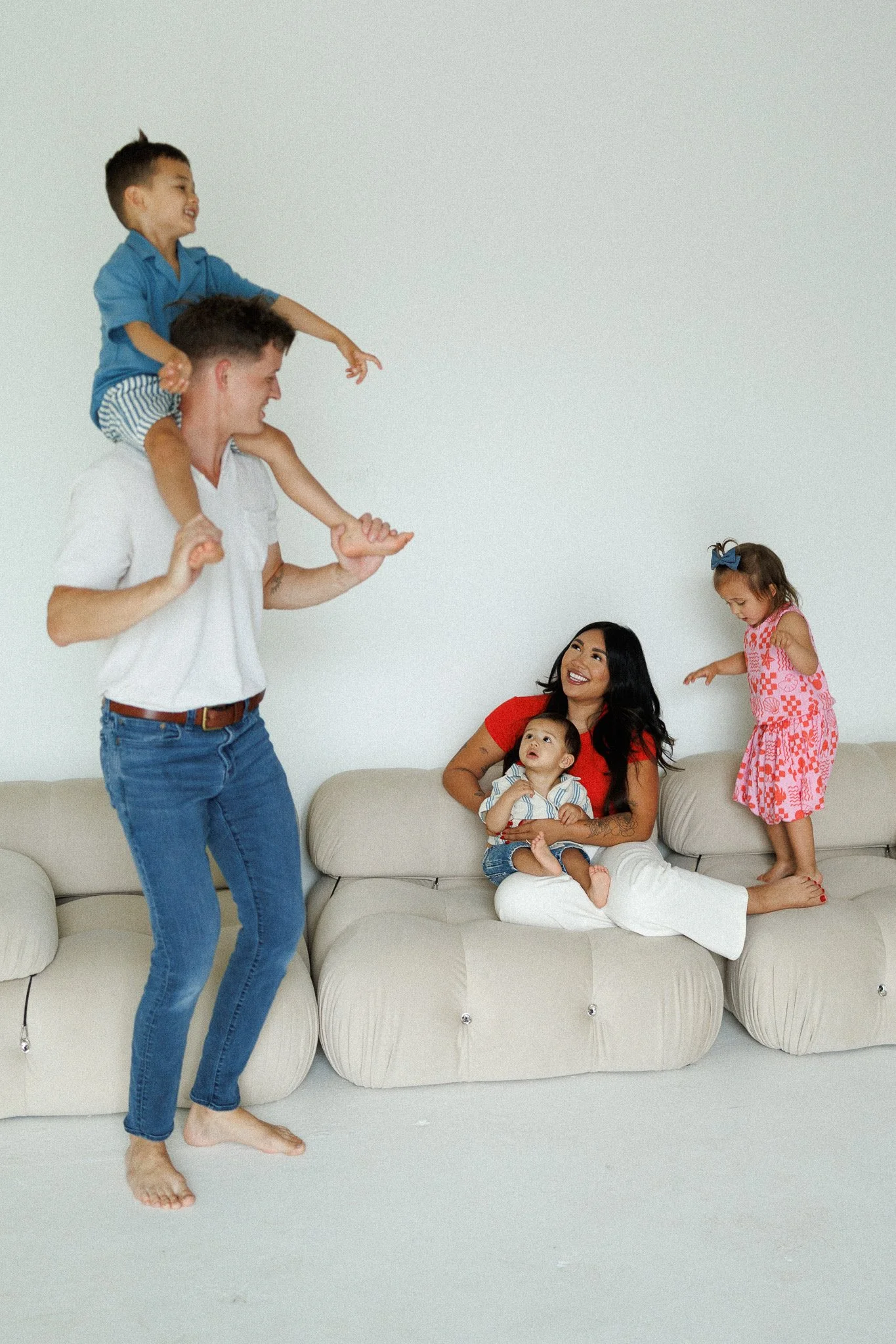A man playing with children, including two boys and two girls, in a living room with a white wall and beige sofa. The man is smiling and has a boy on his shoulders, a girl standing on the sofa, and another boy and girl sitting or sitting nearby.