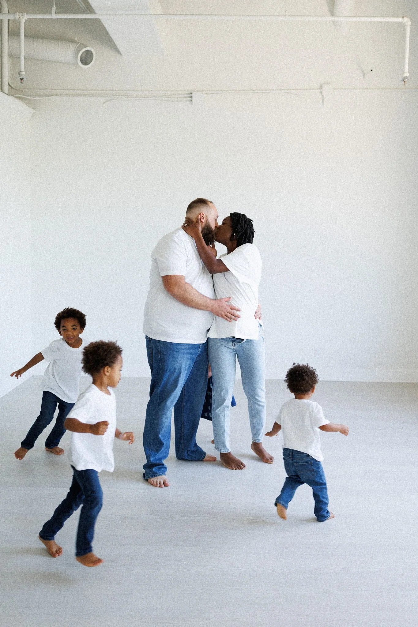 A diverse family sharing a kiss in a white room with five children playing around them.