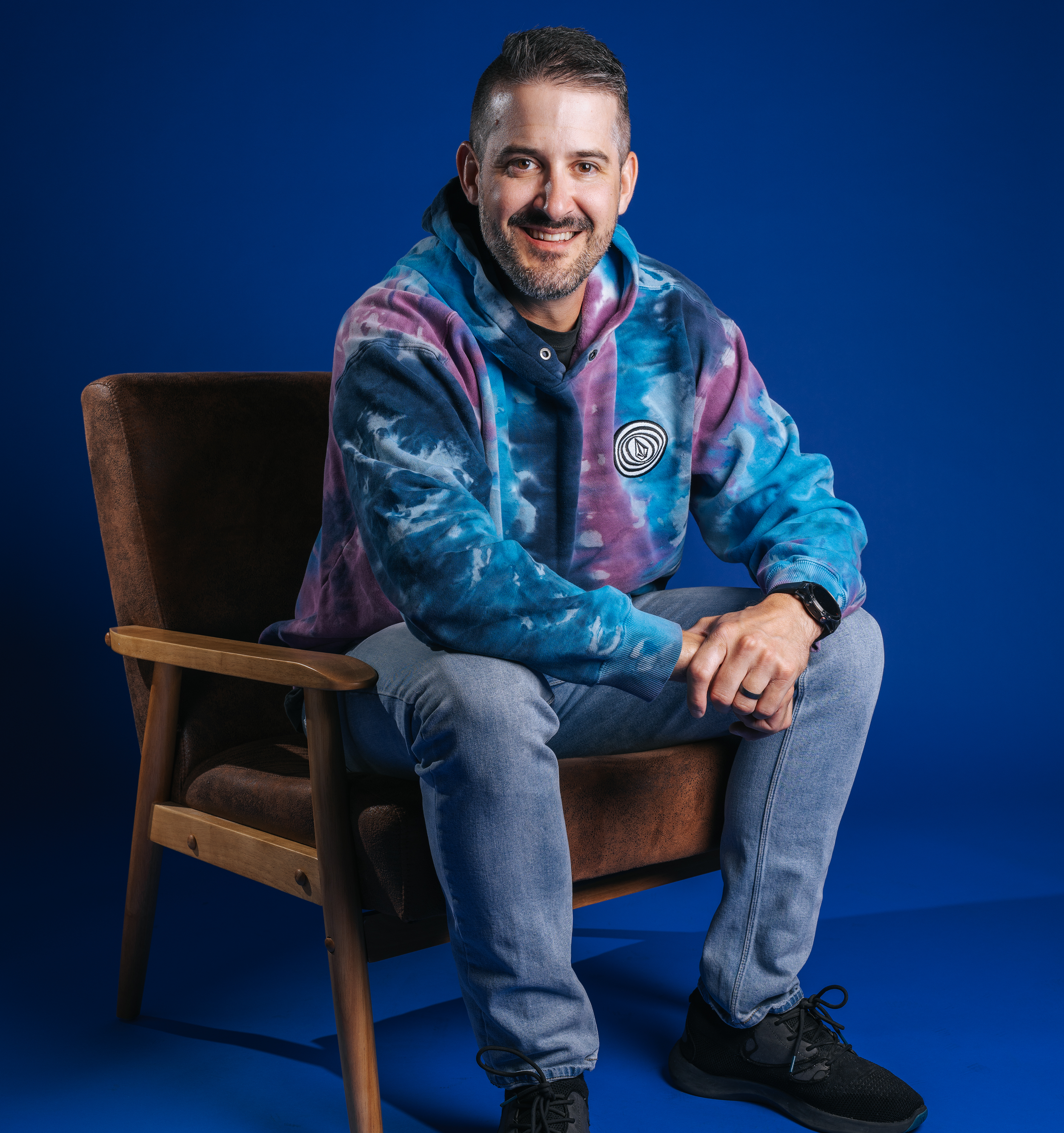 A smiling man sitting on a wooden chair with a brown cushion and backrest against a solid blue background. He is wearing a multicolored tie-dye hoodie, light blue jeans, black sneakers, and a black wristwatch.
