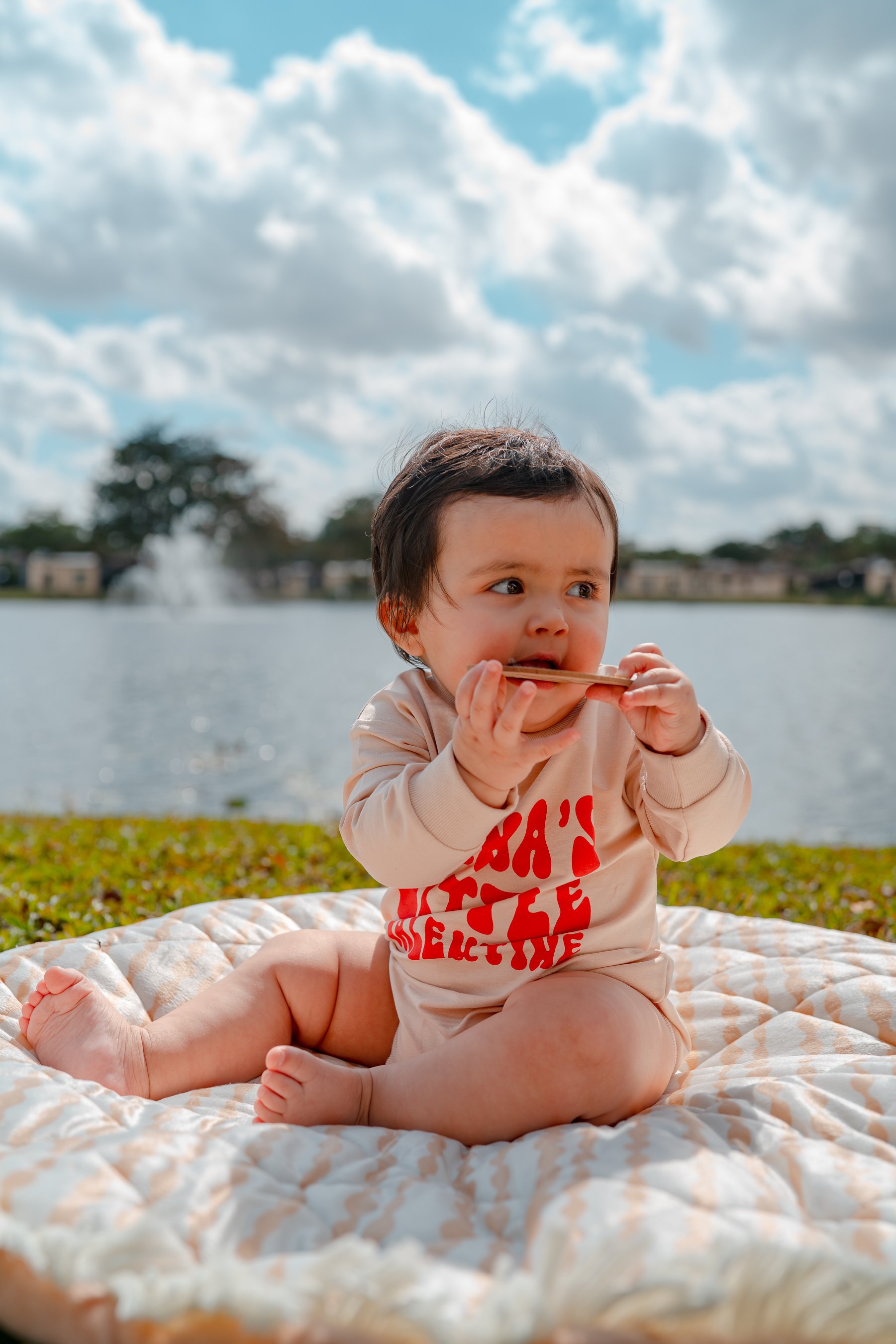 A baby sitting on a quilt outdoors near a lake, chewing on a wooden object, with trees, houses, a fountain, and blue sky with clouds in the background.