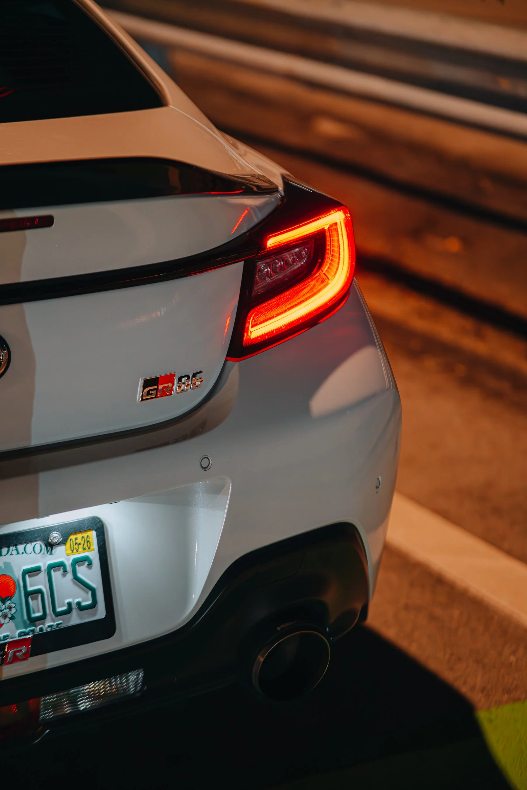 Close-up of the rear end of a silver Toyota GR 86 sports car, showing the taillight illuminated, a license plate, and a exhaust pipe, parked on a street at night.