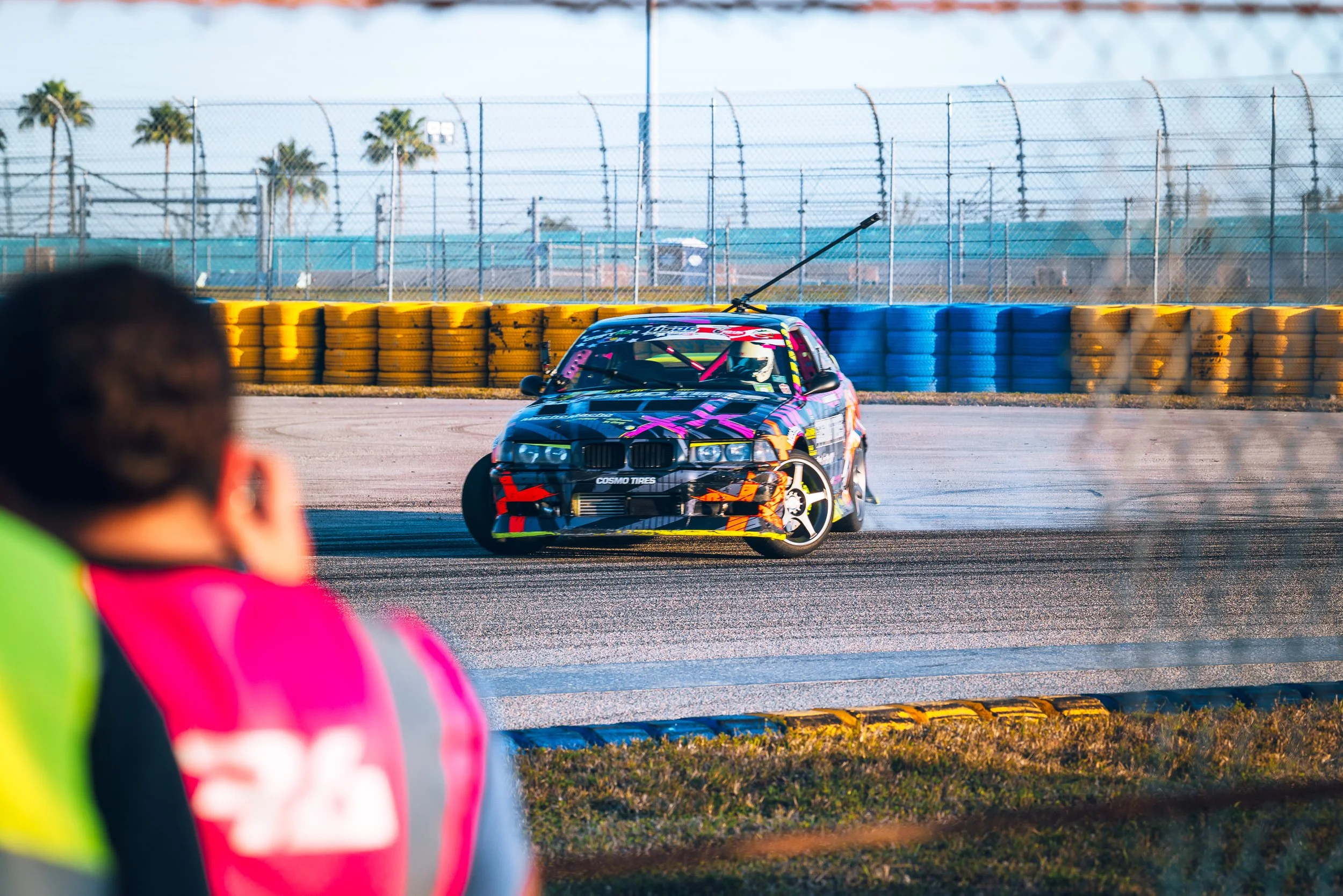 A race car with a colorful, geometric design drifts on a racing track, with a person in a pink vest and blurred foreground watching.