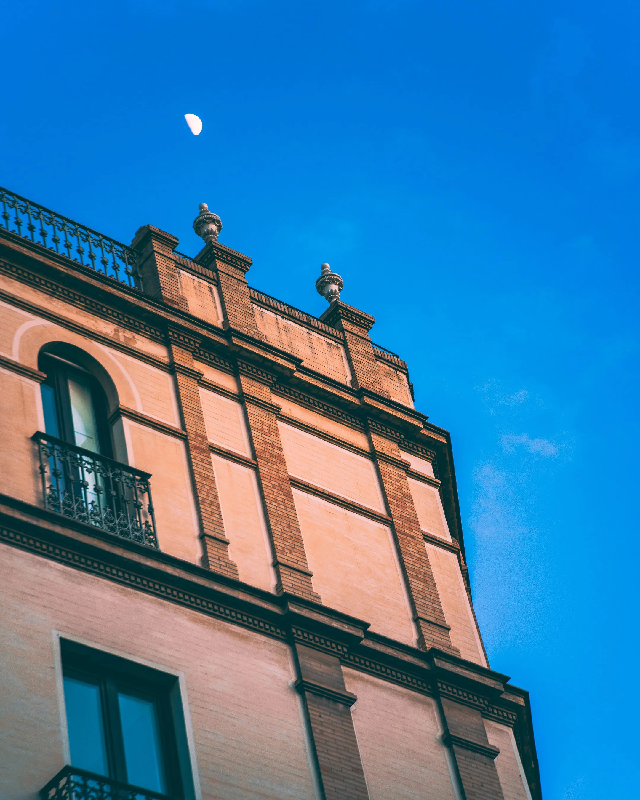An upward view of a historic building with ornate brickwork and wrought iron railings under a blue sky with a crescent moon.