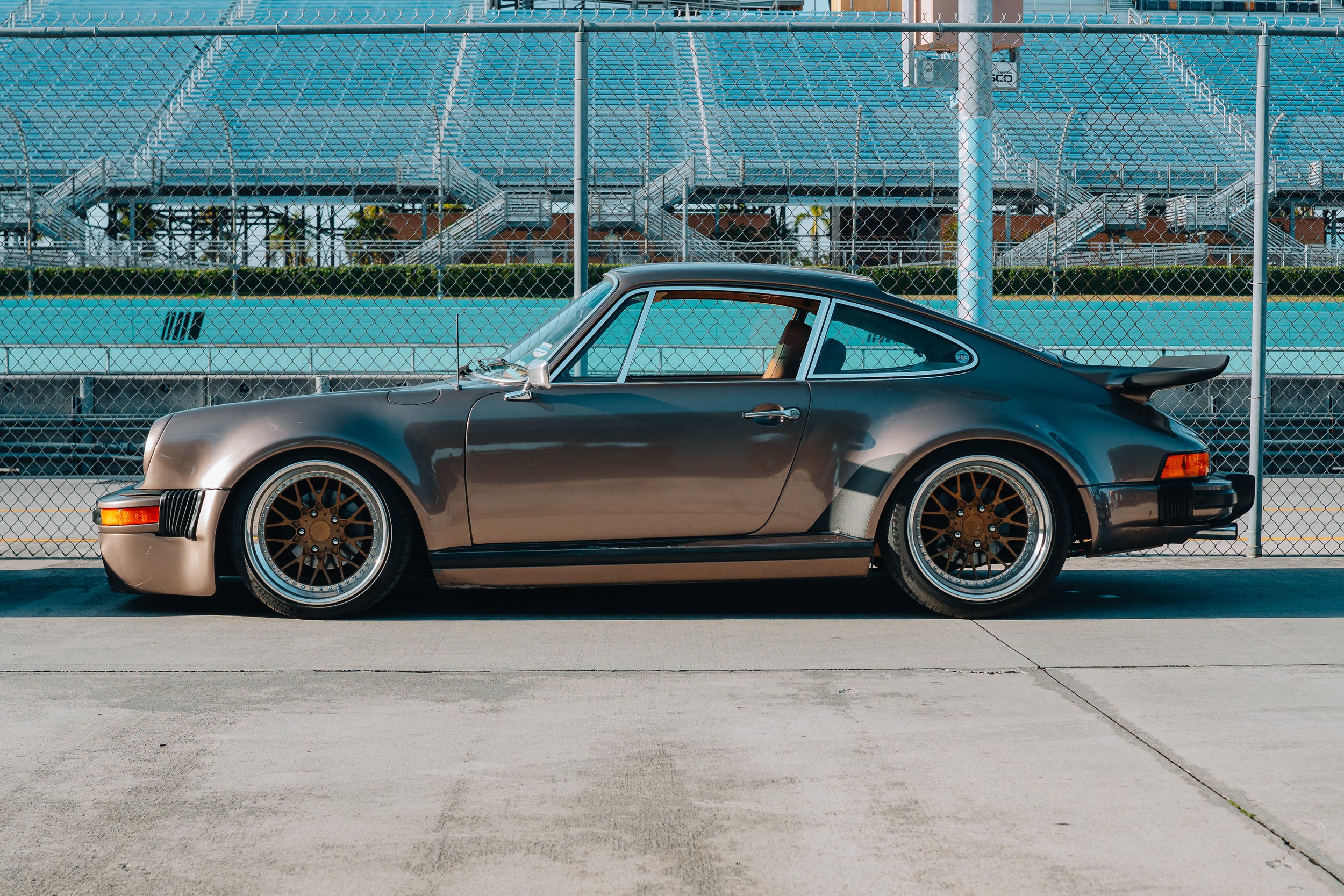 Side view of a vintage black Porsche 911 parked on a concrete surface in front of a chain-link fence with blue bleachers in the background.