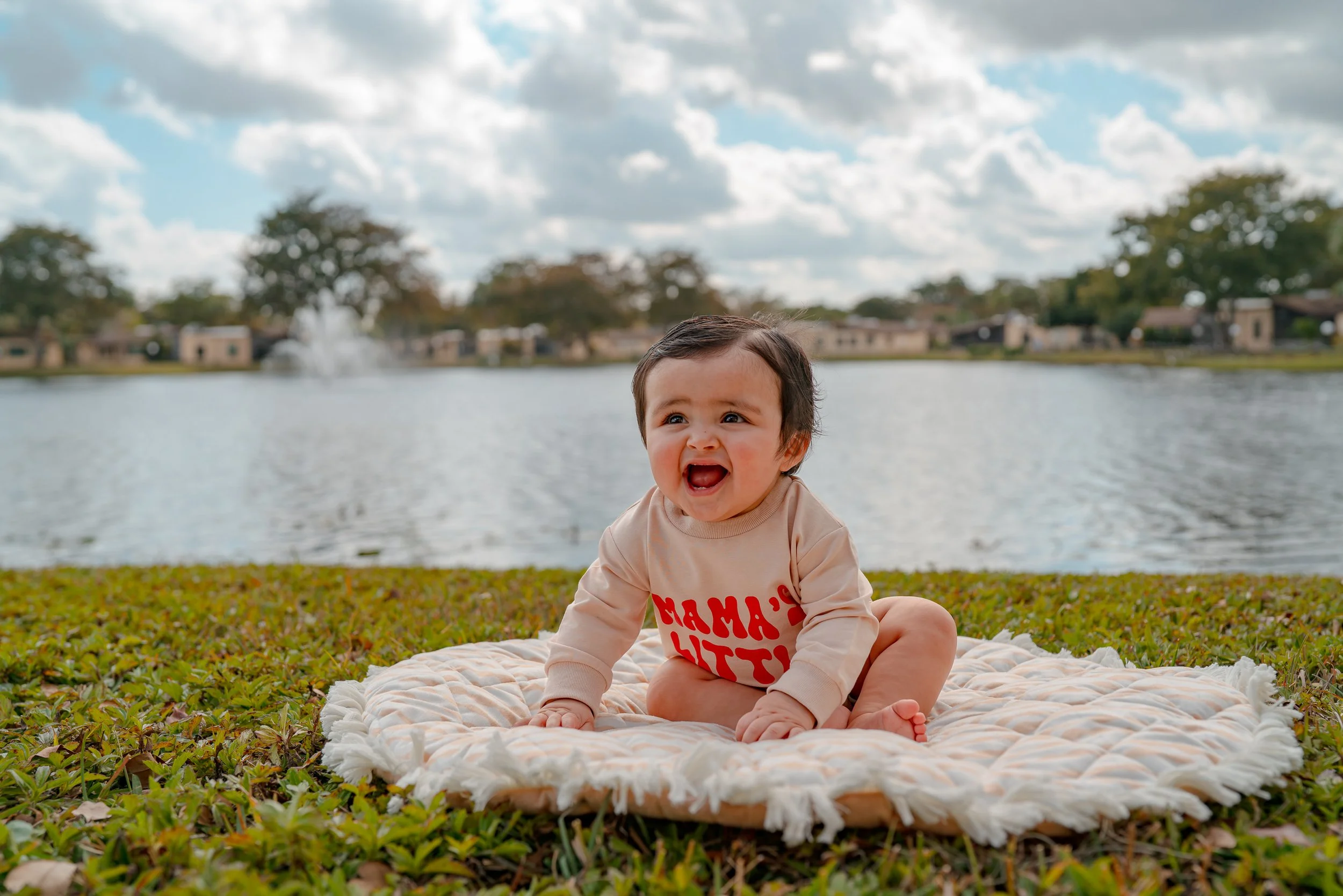 A baby sitting on a blanket near a lake with houses and trees in the background, under a partly cloudy sky.