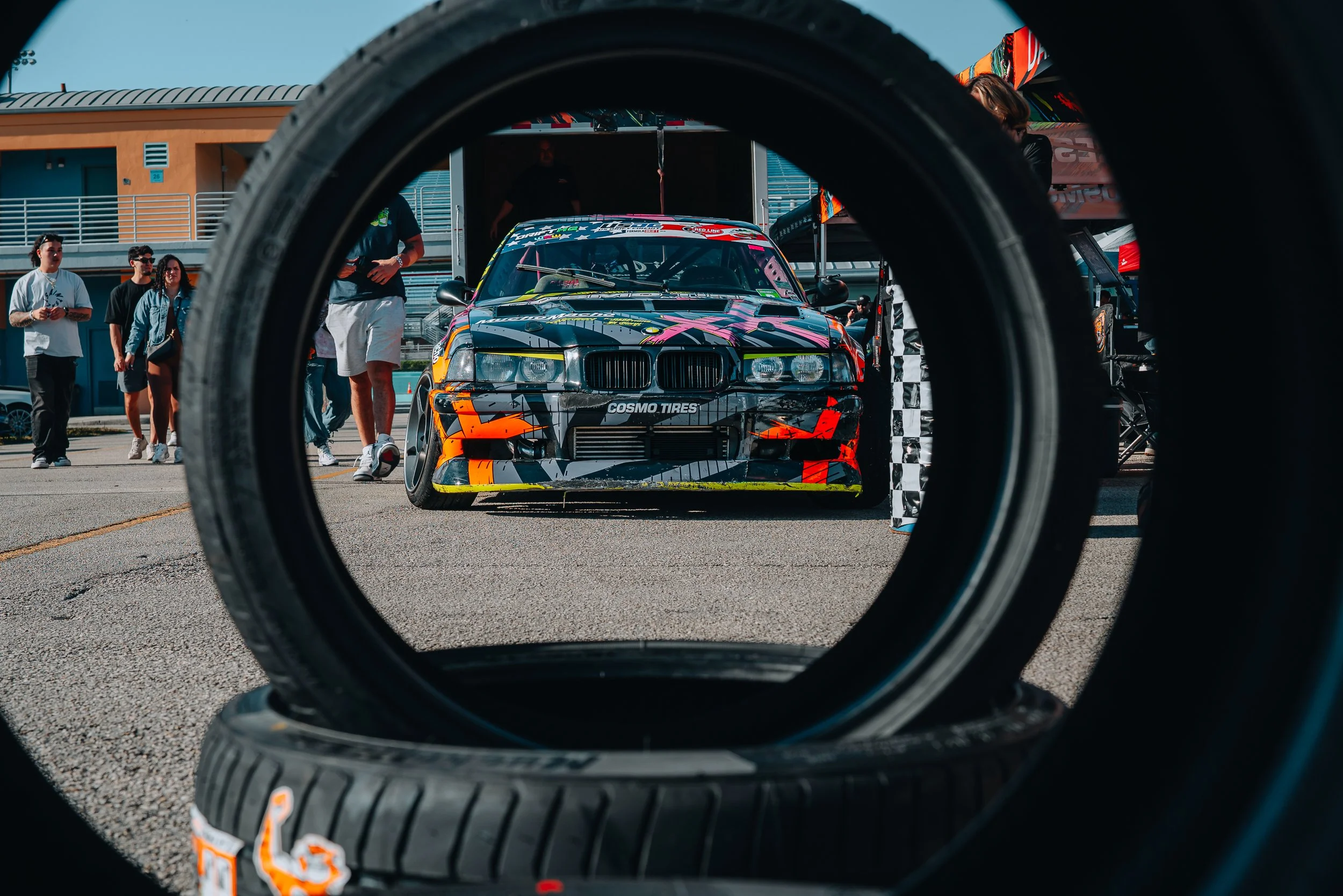 A race car is viewed through two stacked tires on the ground, with people and a pit area in the background at a racing event.