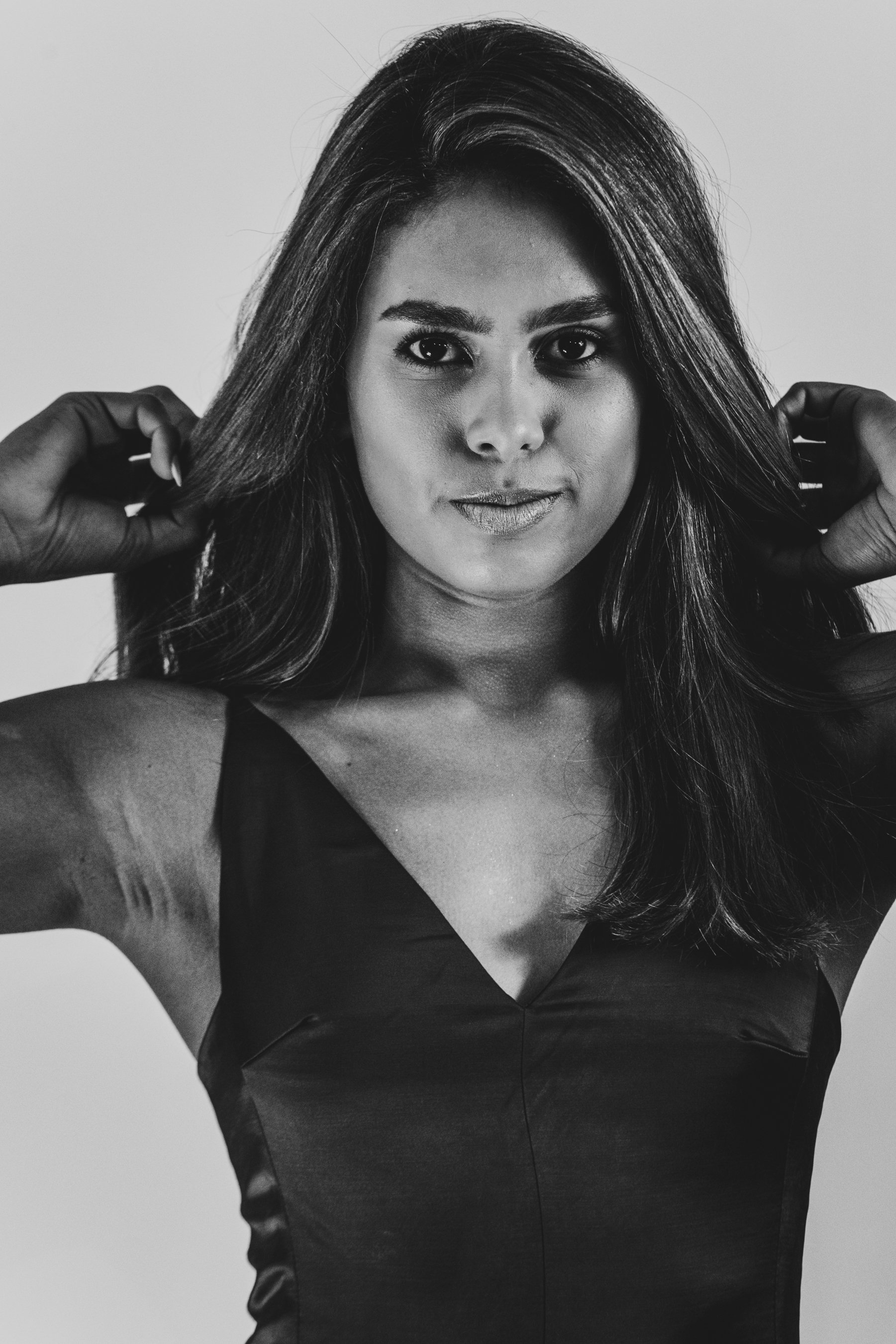 Black and white portrait of a woman with long hair, wearing a sleeveless top, touching her hair with both hands, looking directly at the camera.