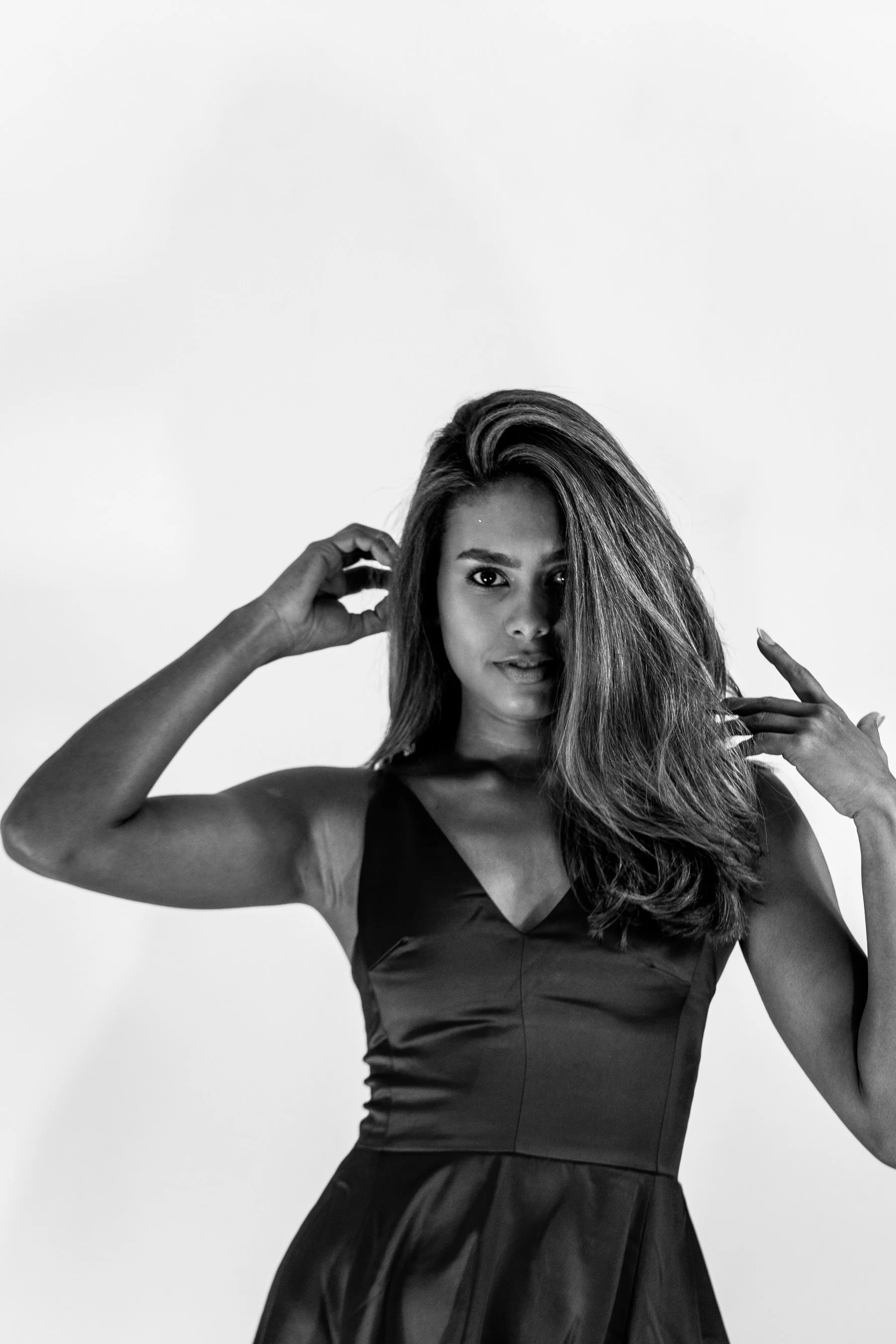 Black and white photo of a woman with long hair, wearing a sleeveless dress, posing with one hand near her head and the other touching her hair.