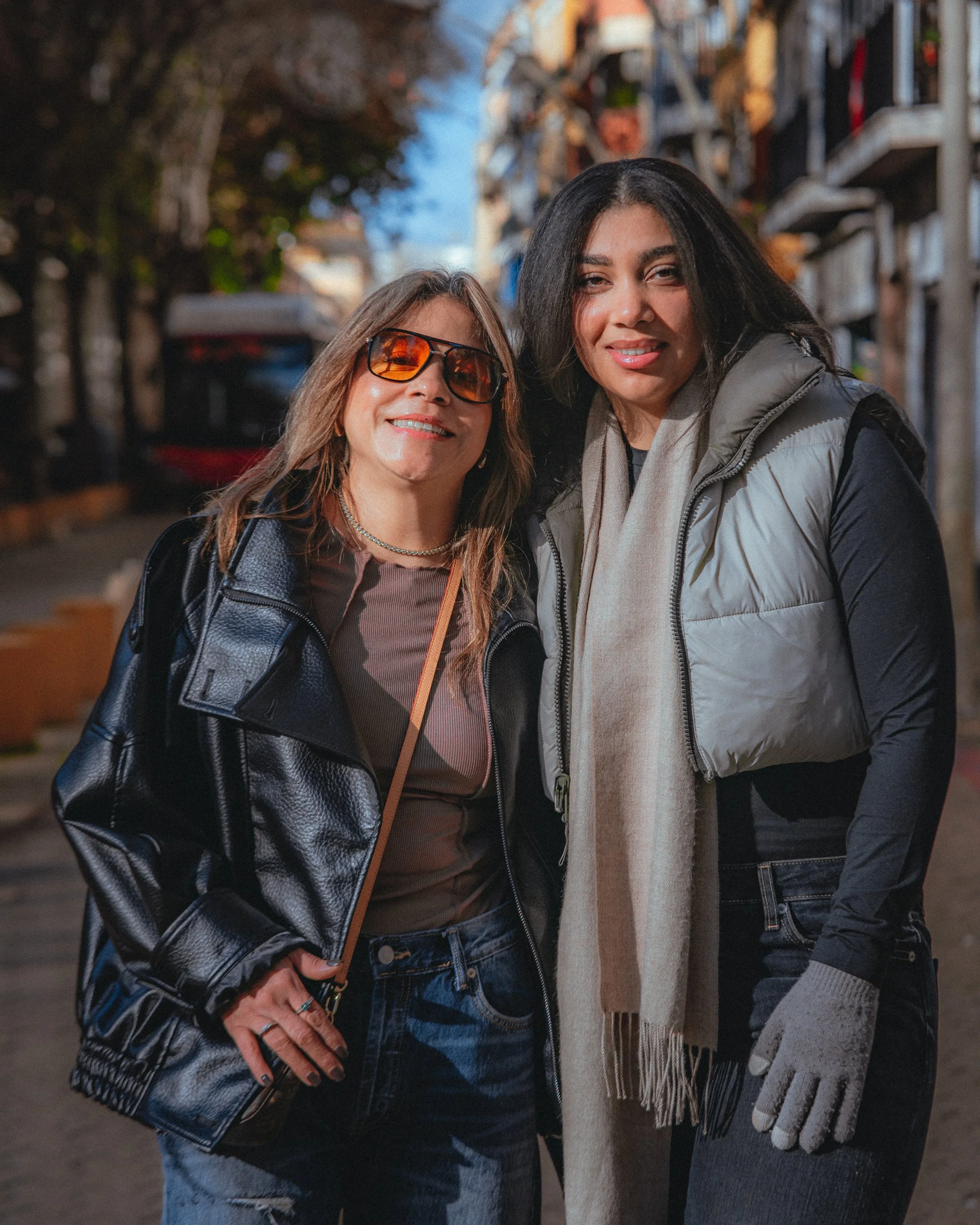 Two women standing close together outdoors on a city street, smiling at the camera, with trees and buildings in the background during daytime.