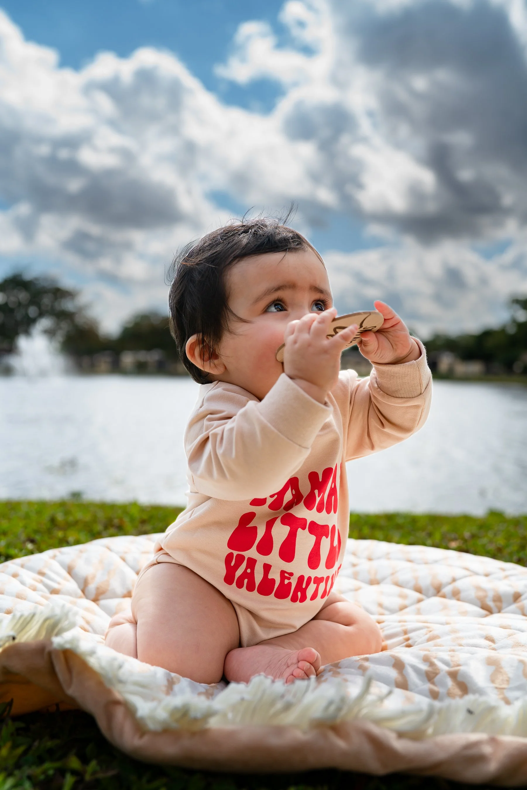 A young child with dark hair sitting on a blanket outdoors near a body of water, holding a wooden spoon to her nose, with a cloudy sky in the background.