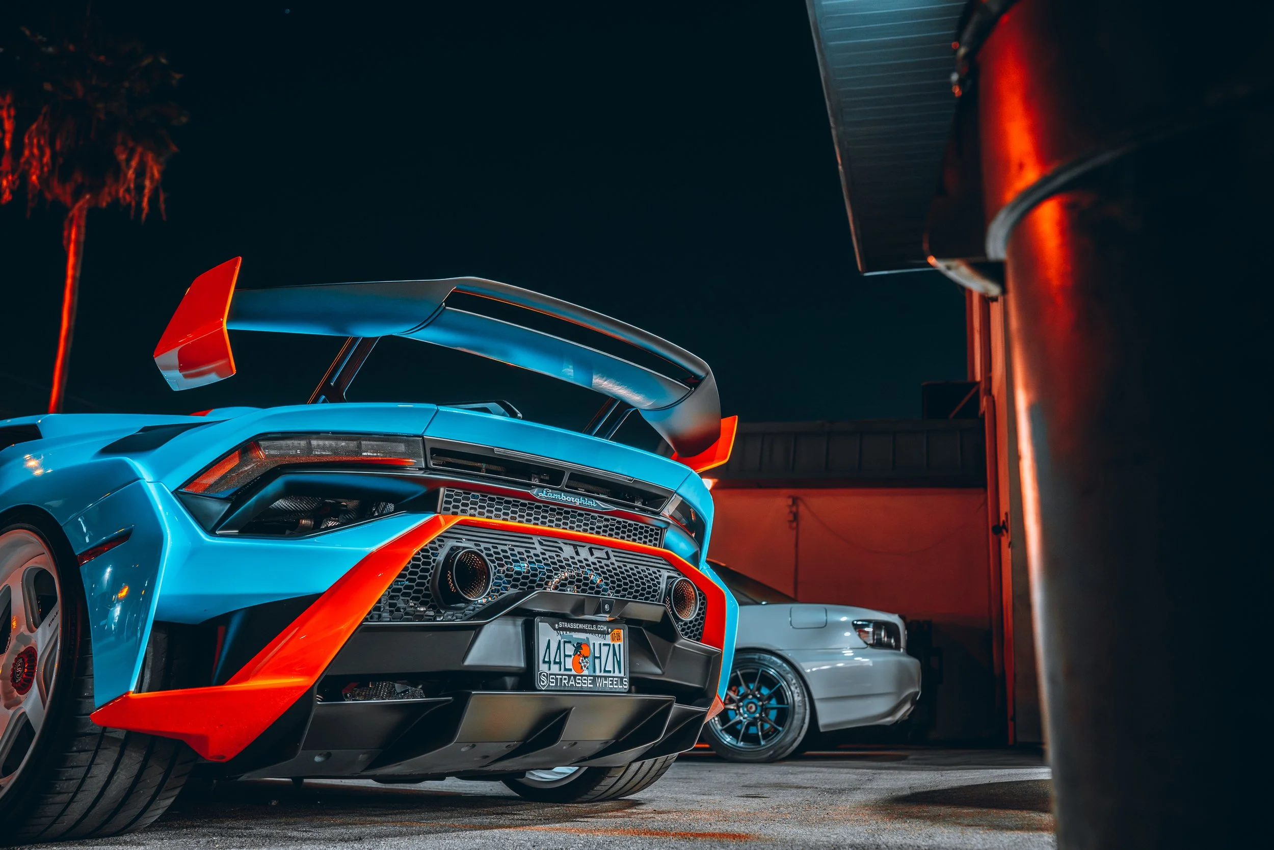 A blue Lamborghini sports car with orange accents, rear spoiler, and rear diffuser, parked at night in a garage with another silver sports car in the background. A palm tree is visible to the left, and a large metallic object is on the right.