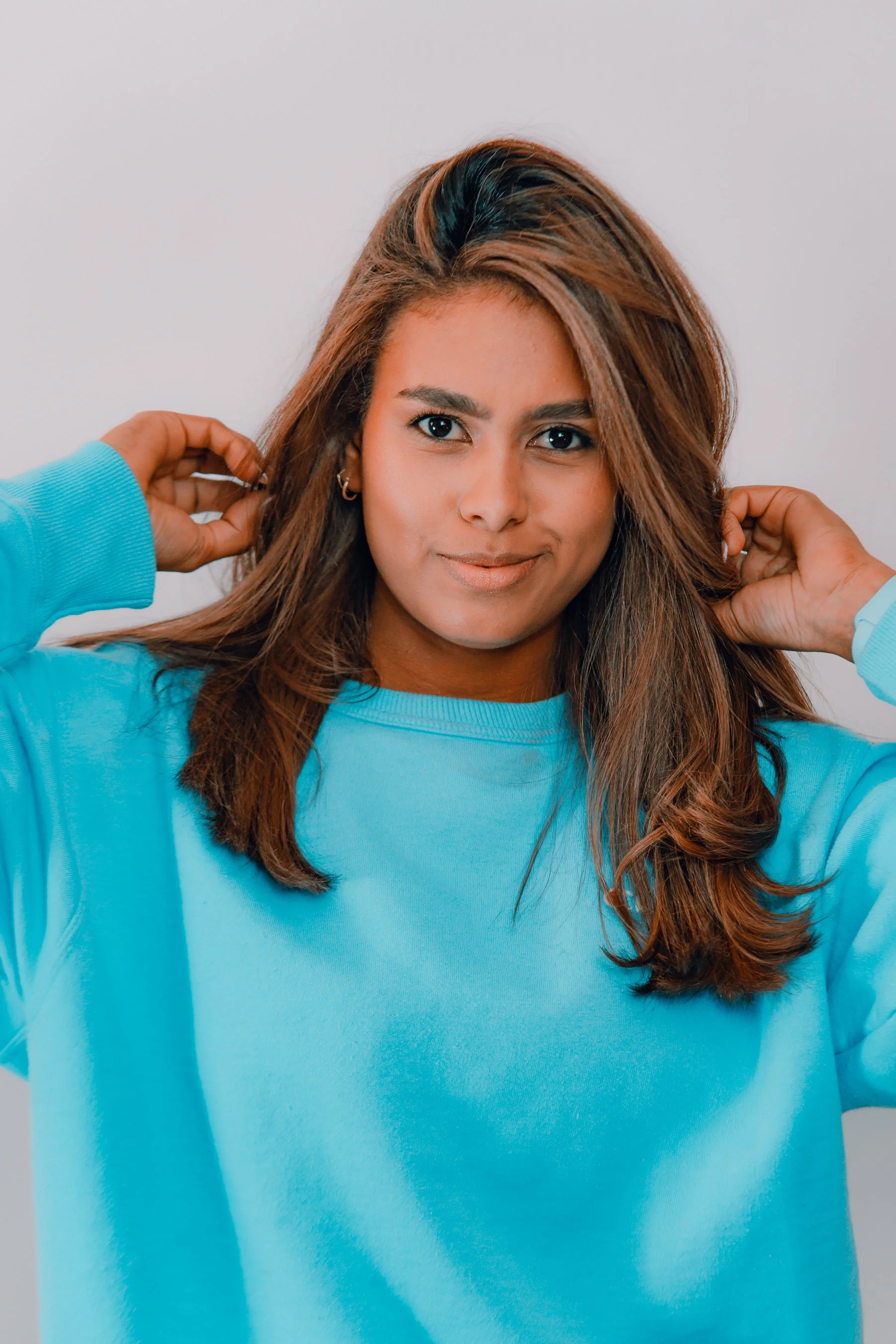 A woman with long brown hair and earrings, wearing a light blue sweatshirt, looking at the camera with a slight smile, touching her hair.